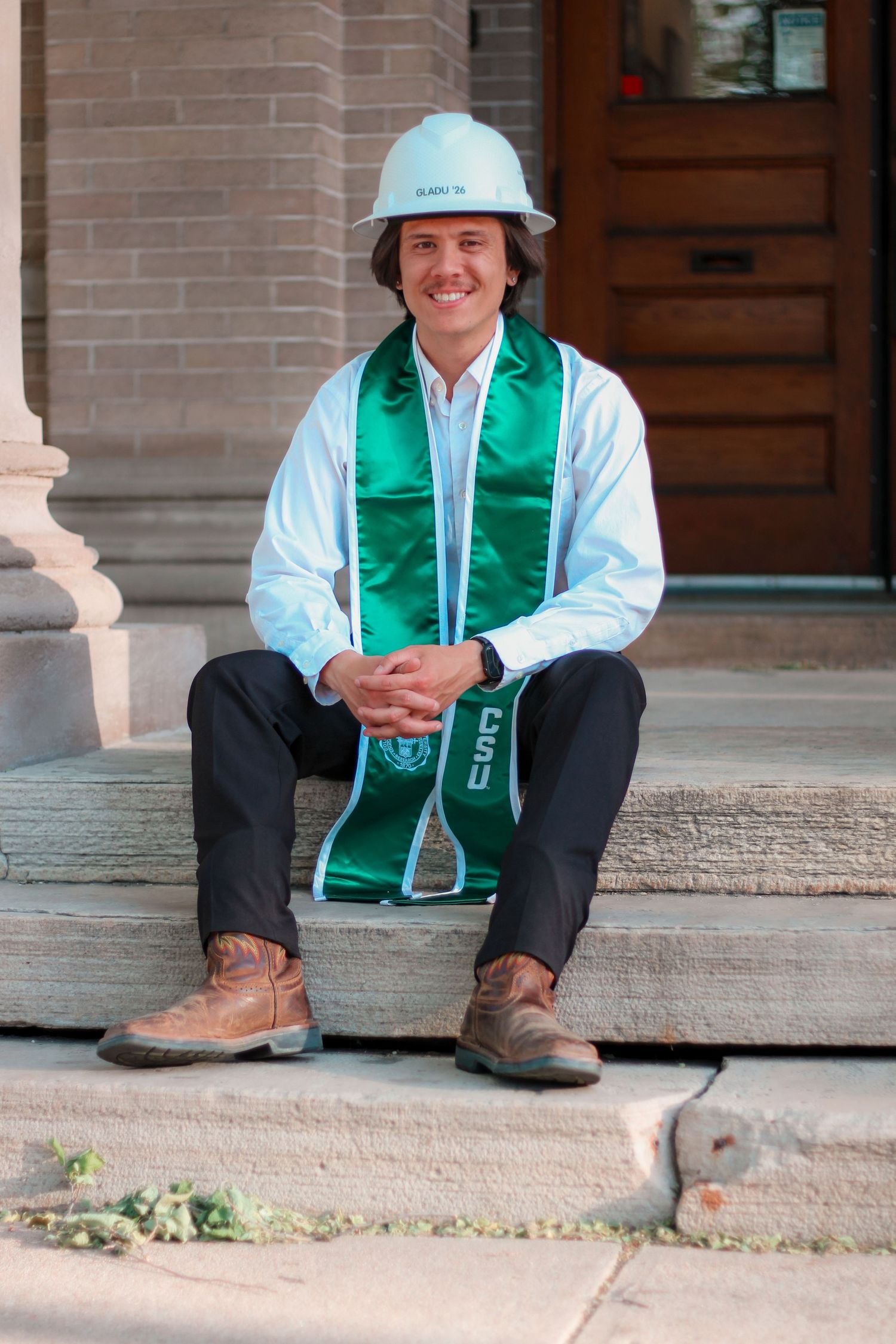 Smiling college graduate sitting on steps wearing a green CSU stole and white hard hat outdoors.
