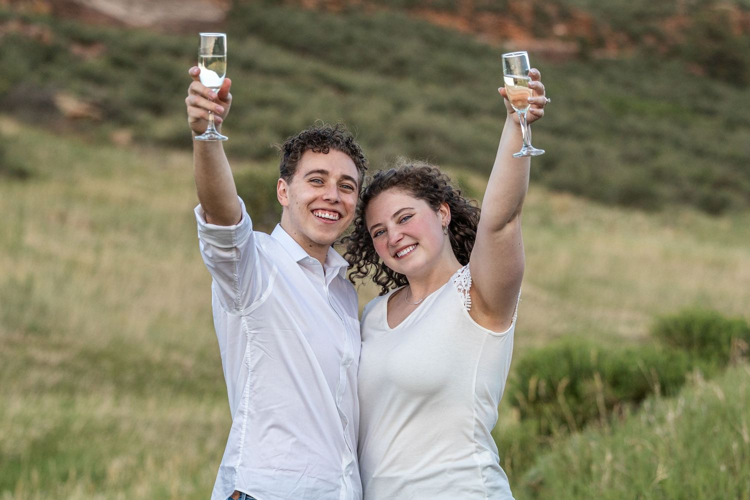 Happy couple in white attire toasting with champagne glasses outdoors in a lush green landscape.