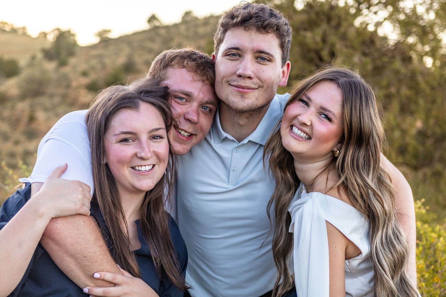 Four young adults smiling and posing together outdoors with green hills in the background.