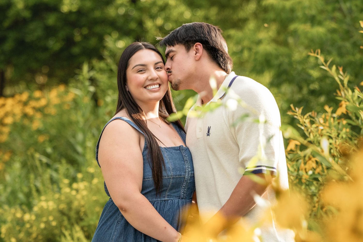 A couple shares a tender kiss outdoors surrounded by lush green foliage and yellow wildflowers.