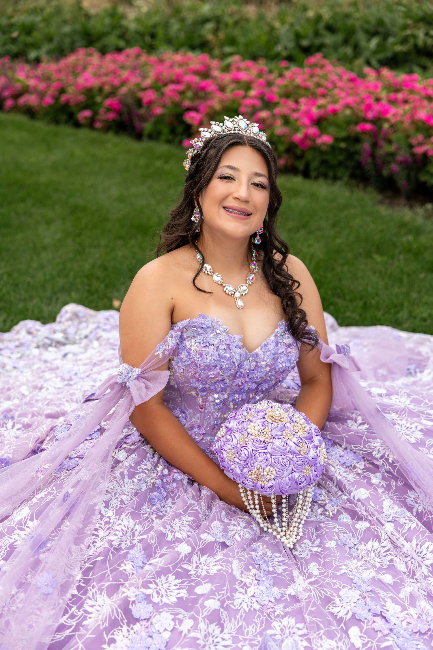Young woman in lavender quinceañera ball gown with tiara and pearl bouquet poses in garden with pink roses.