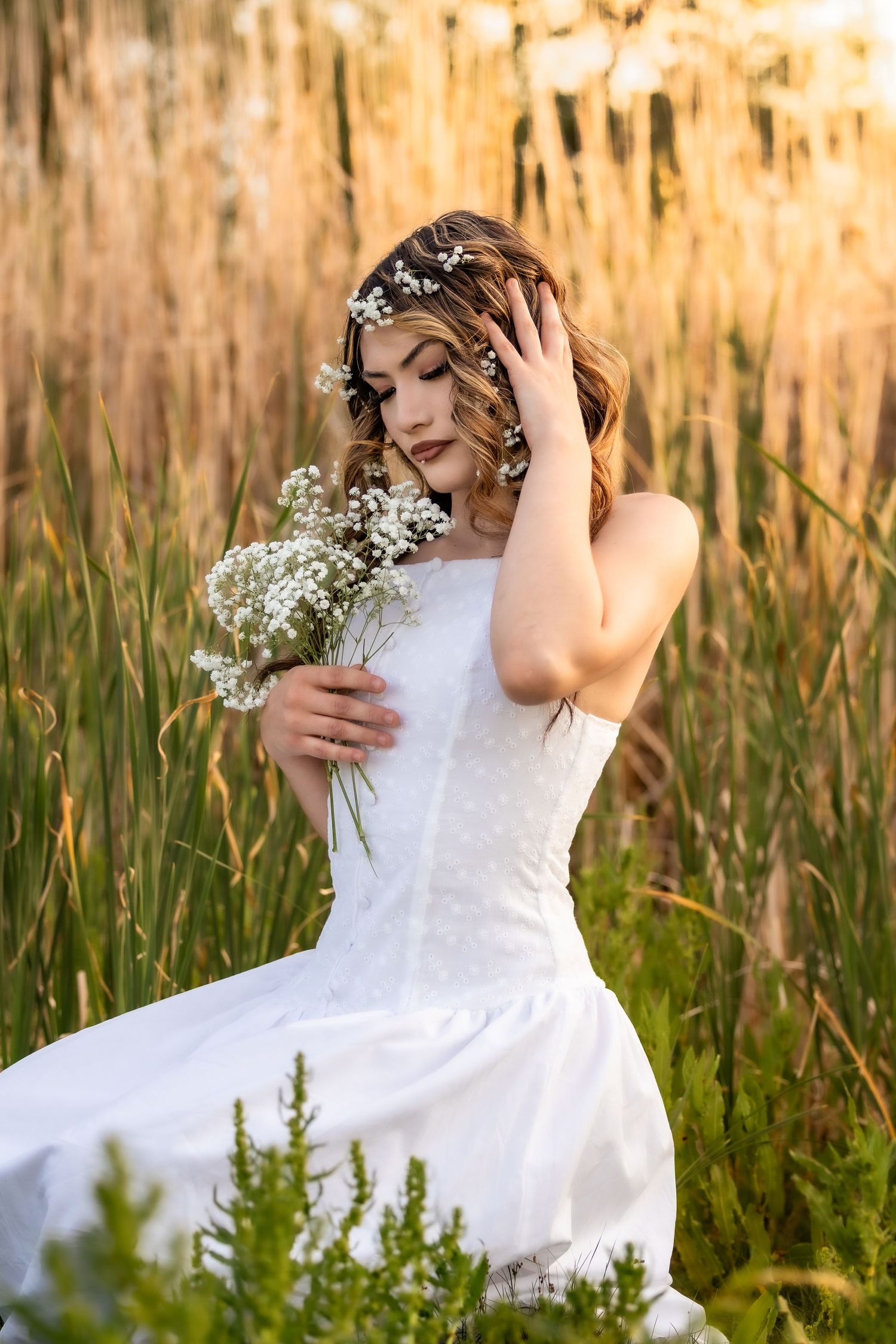 Young woman in white bridal gown holding white wildflowers among tall golden reeds at sunset in Littleton, Colorado.