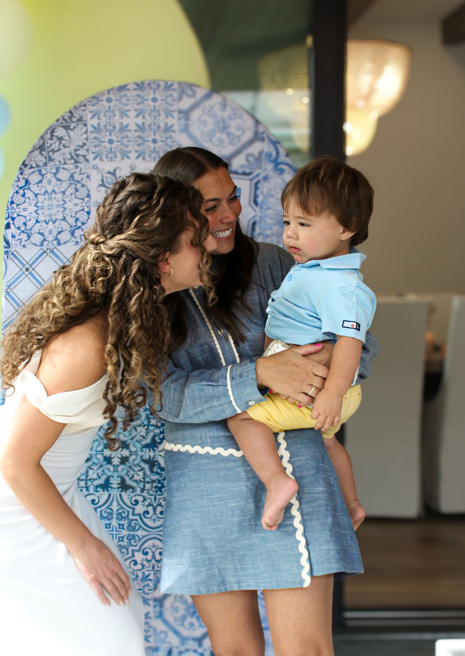 Two women laughing together while one holds a baby boy at a festive blue party event.
