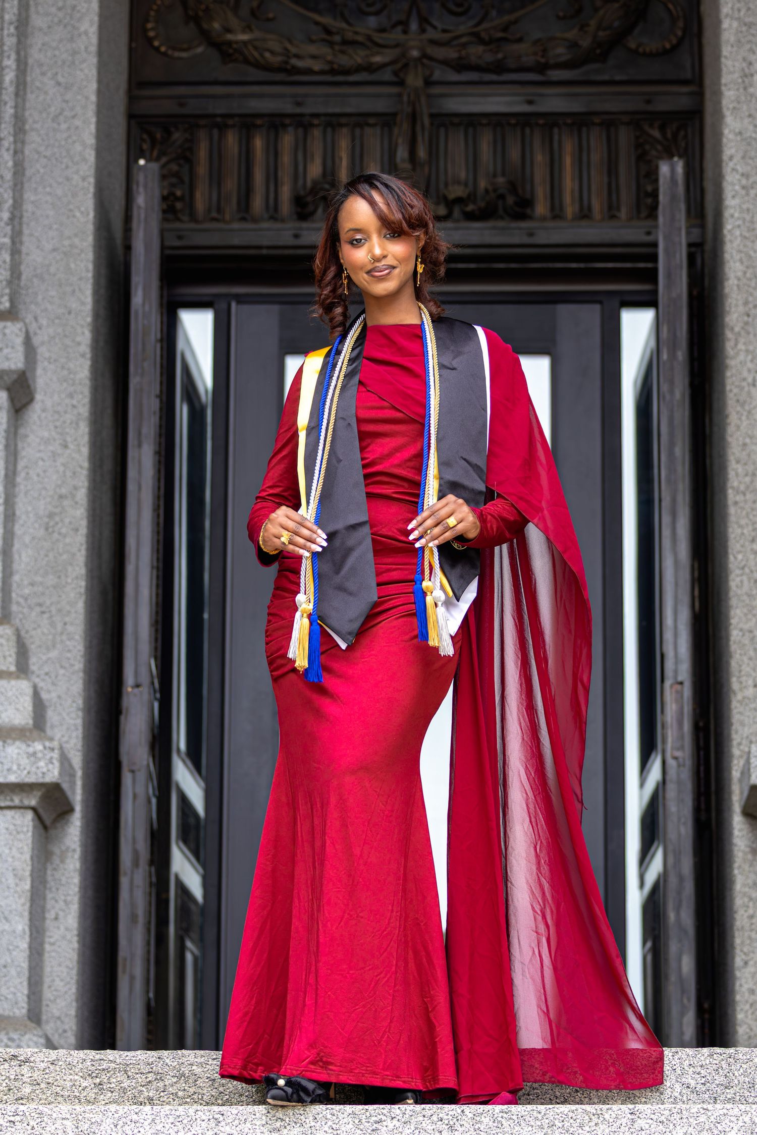 A young woman in a red gown and graduation stoles poses proudly on steps before ornate doors.