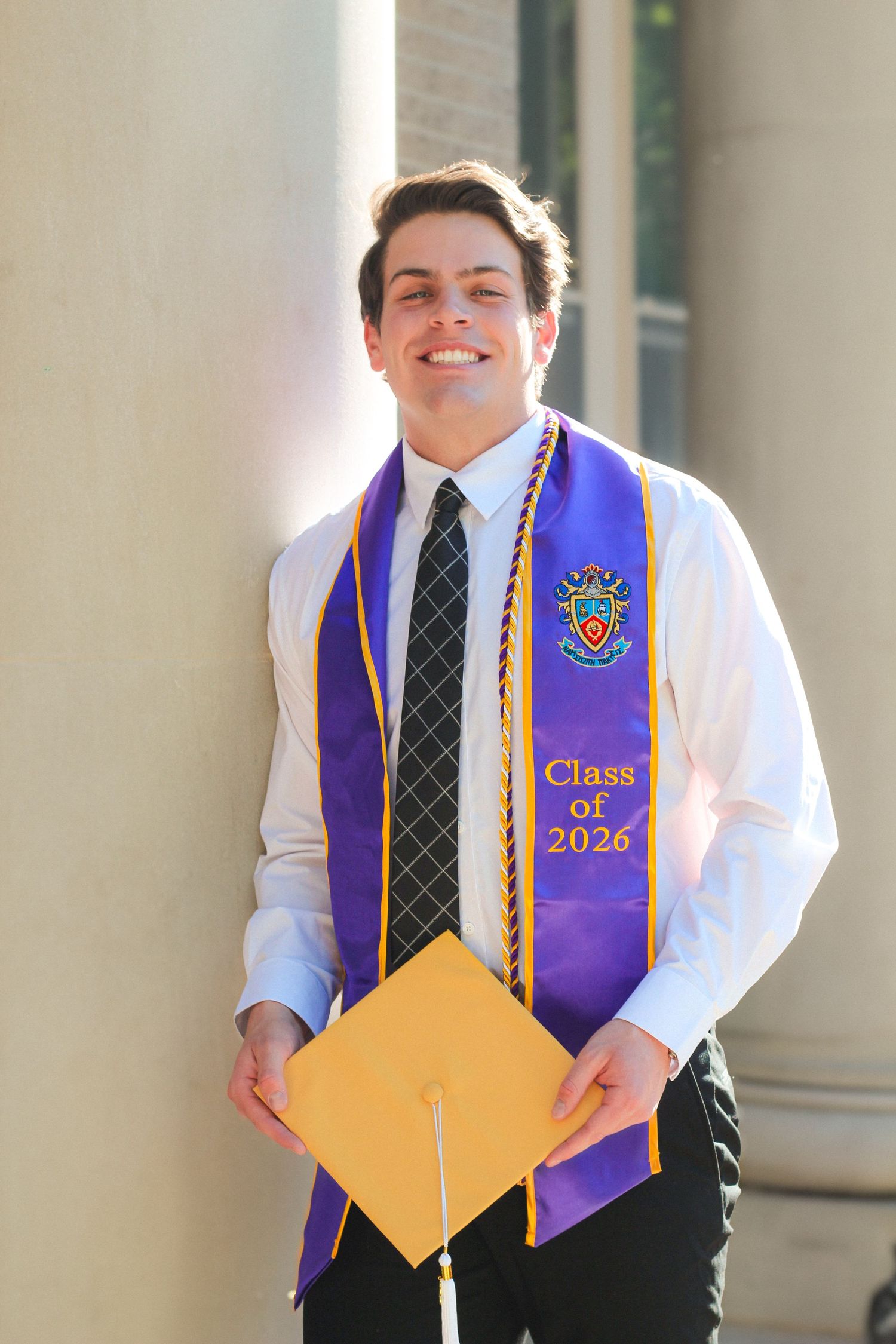 Smiling young man in white shirt and tie wearing purple Class of 2026 graduation stole holding gold cap outdoors.