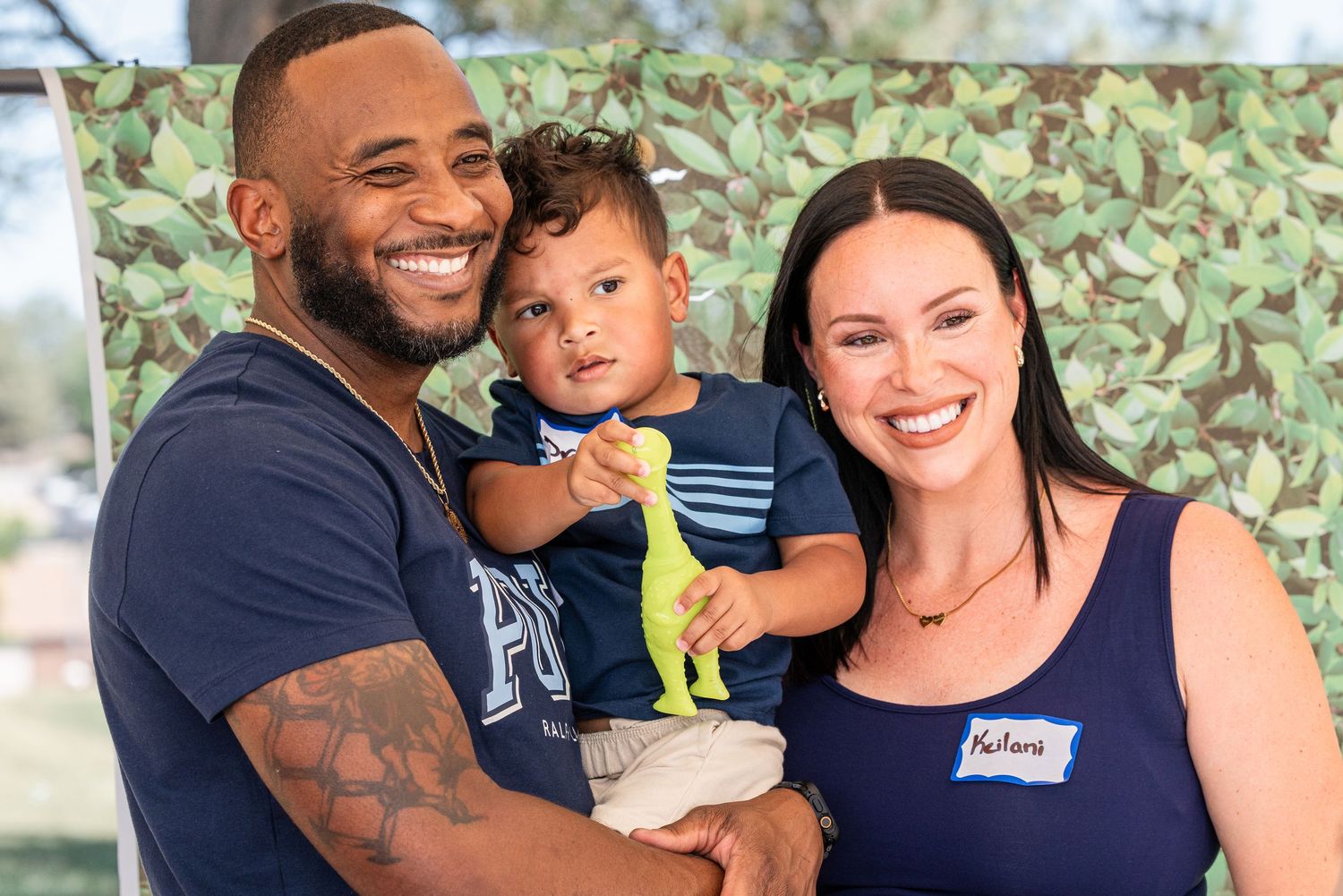 A smiling family with a man, woman, and toddler holding a toy dinosaur posing outdoors at a sunny event.