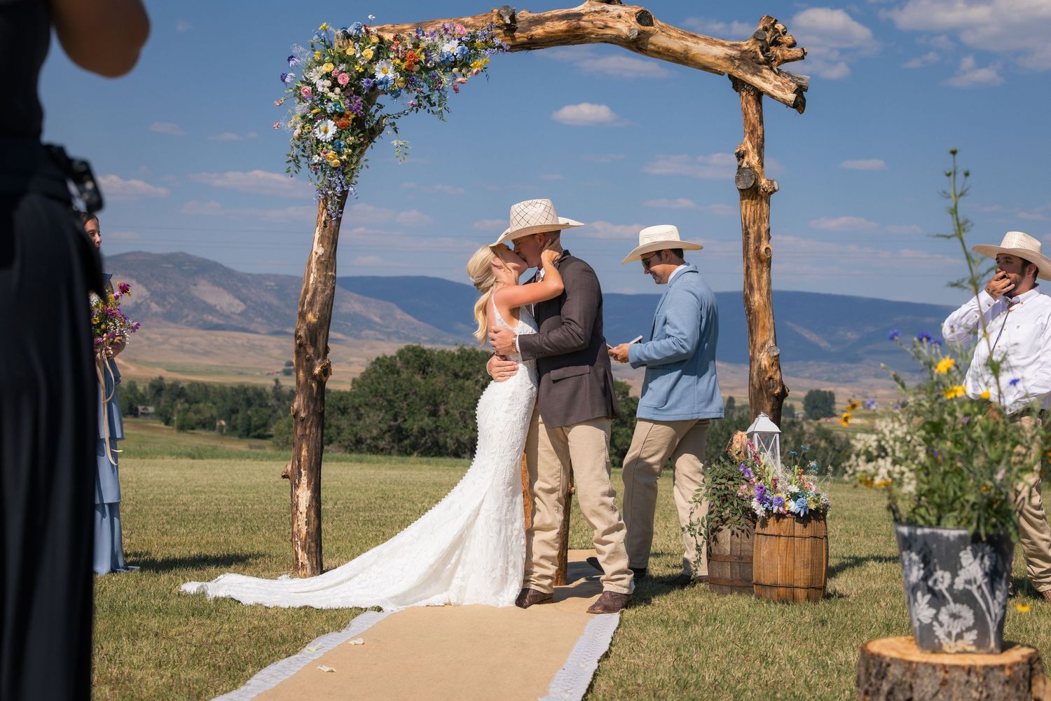 Outdoor wedding ceremony under rustic wooden arch with mountain views, bride and groom kissing in sunny field.