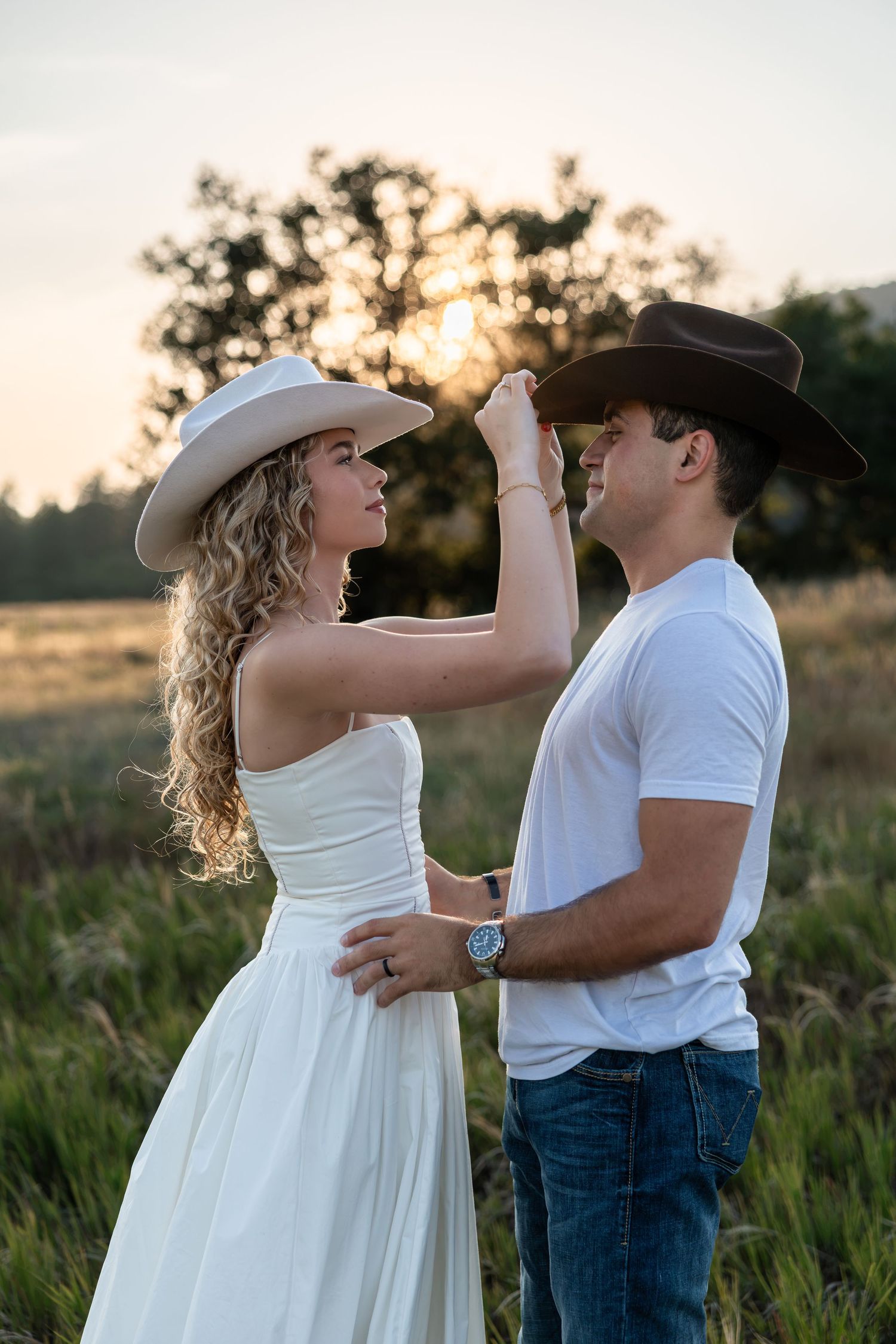 Couple in cowboy hats dancing in a field at sunset, woman in white dress touching man's hat.