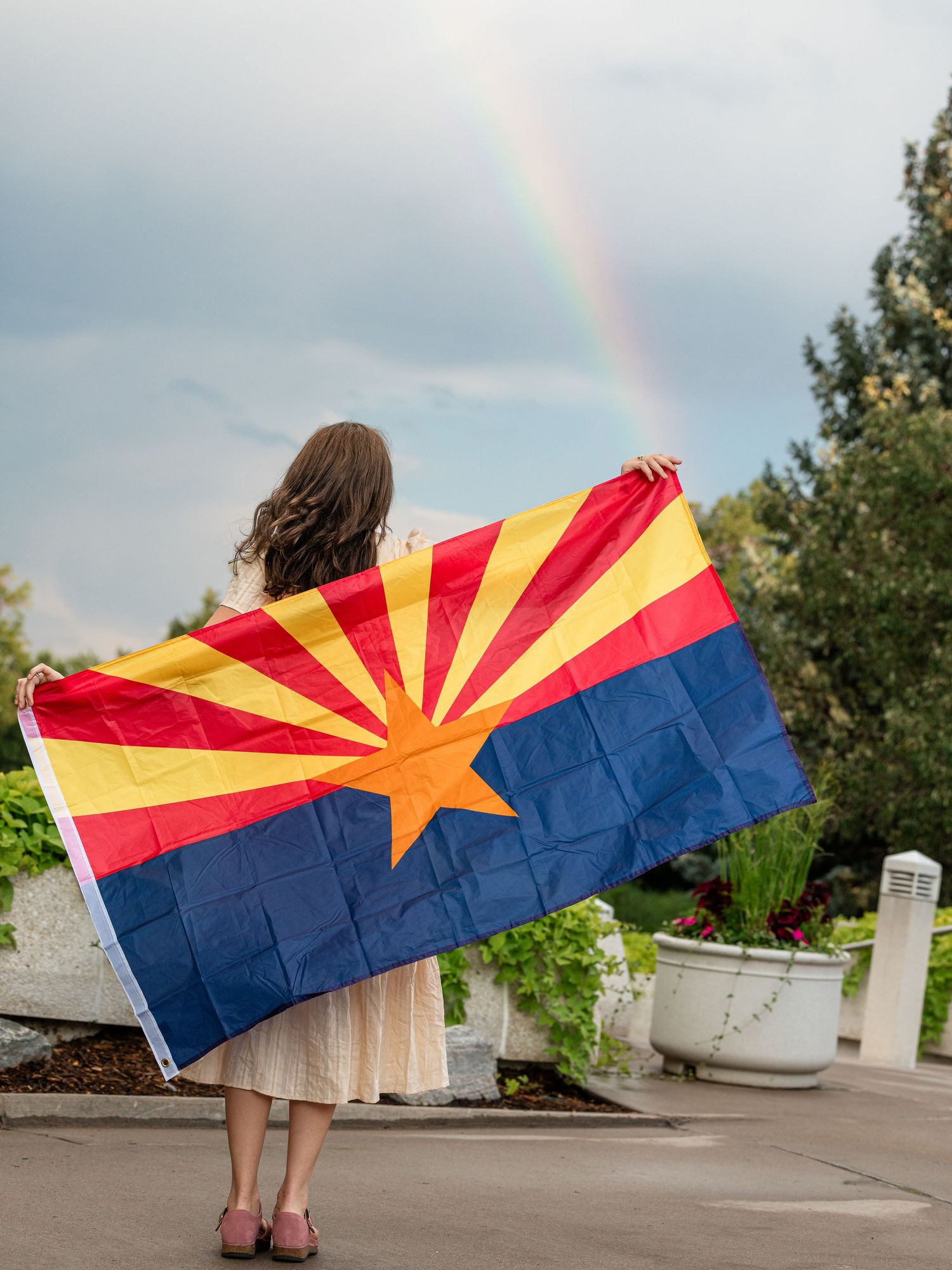 Woman holding Arizona state flag with rainbow in background outdoors.