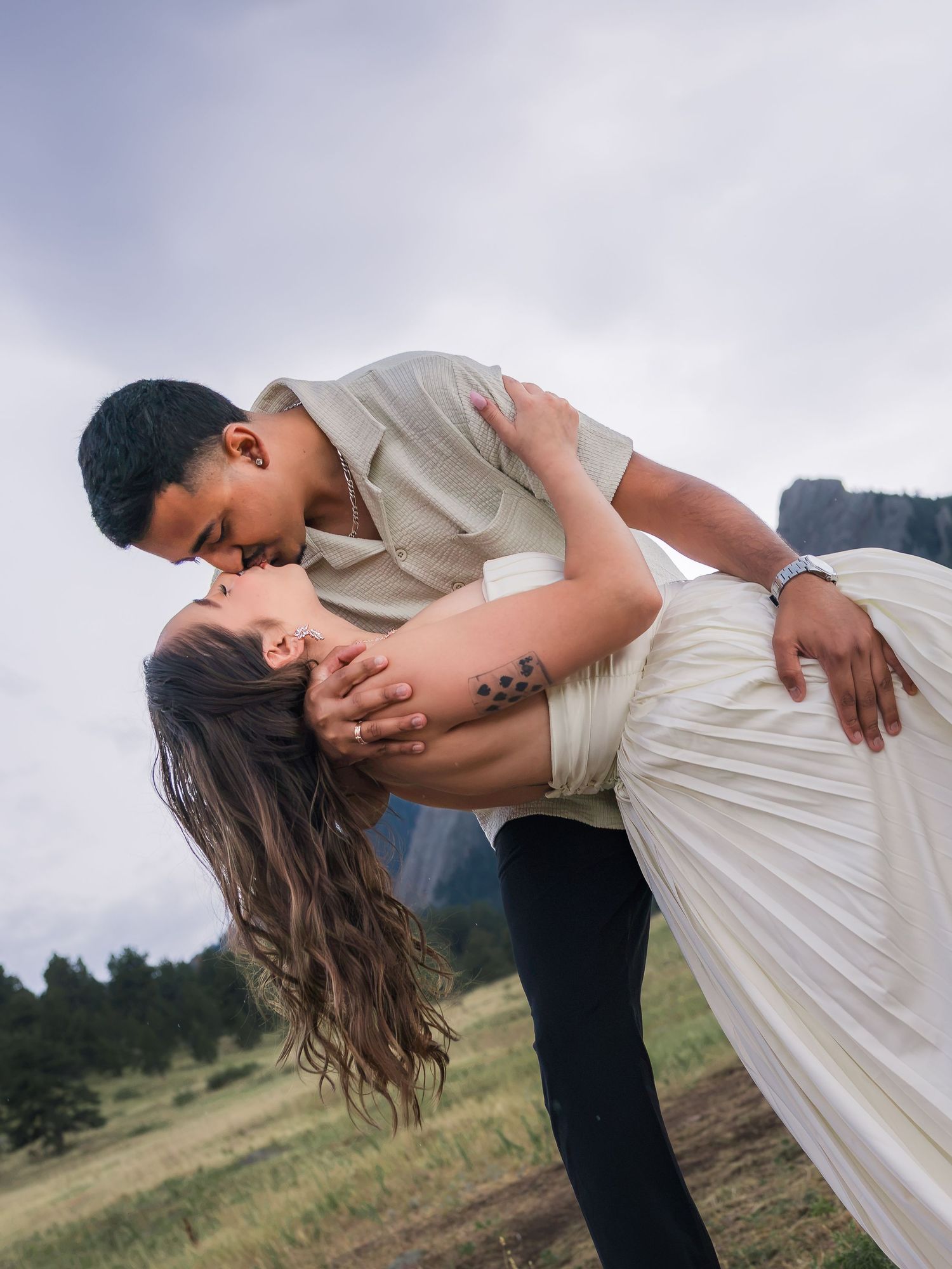 A couple shares a romantic dip kiss in a scenic mountain meadow, the woman in a white dress, outdoors.
