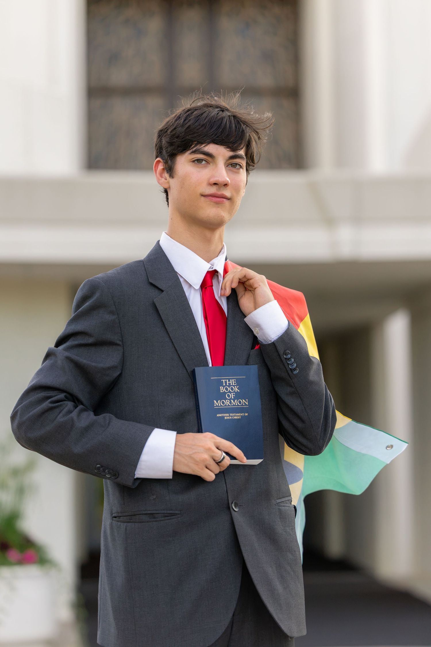 Young man in gray suit holding Book of Mormon and rainbow flag outside a building.