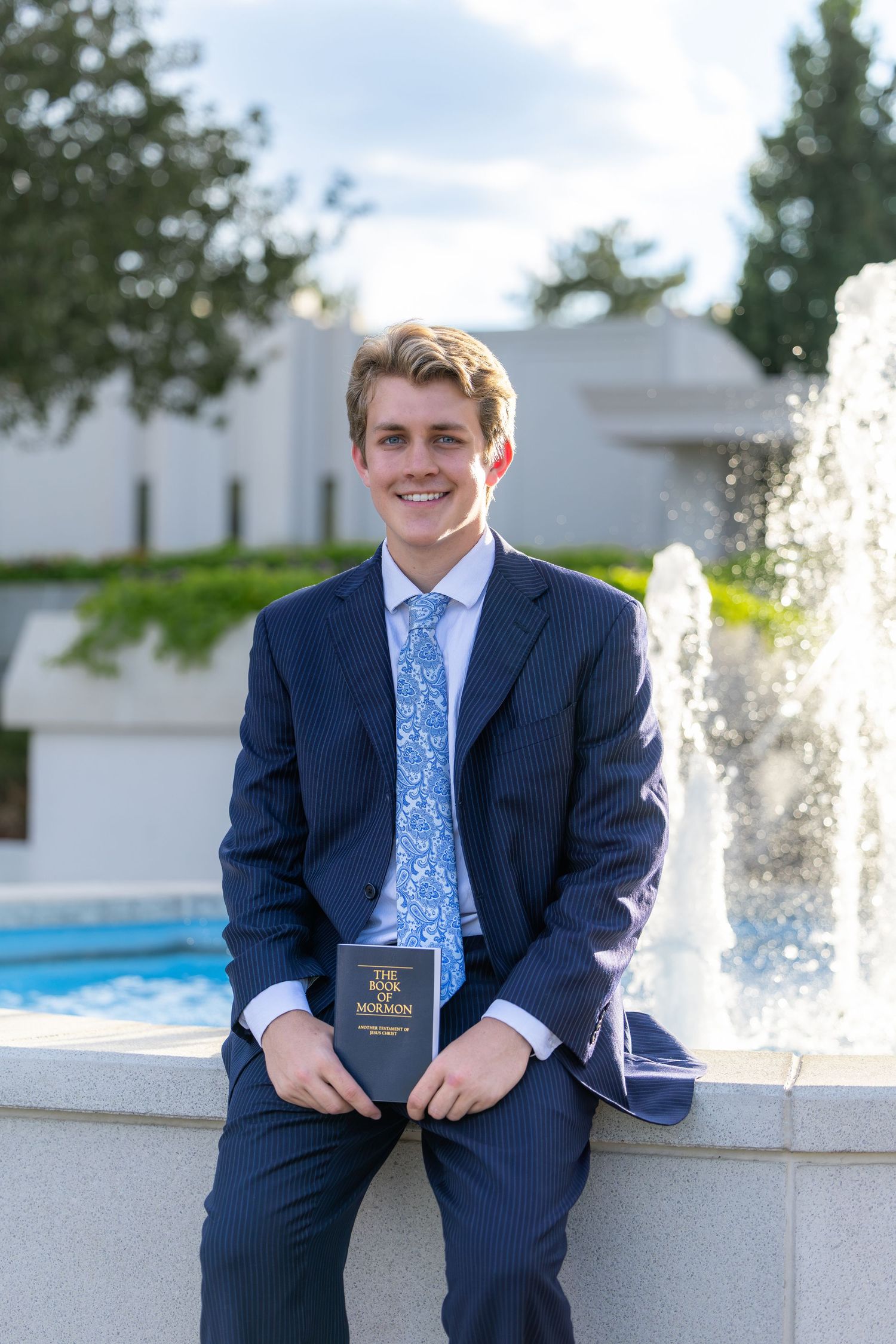 Young man in navy suit holding Book of Mormon sits smiling by a fountain outdoors.