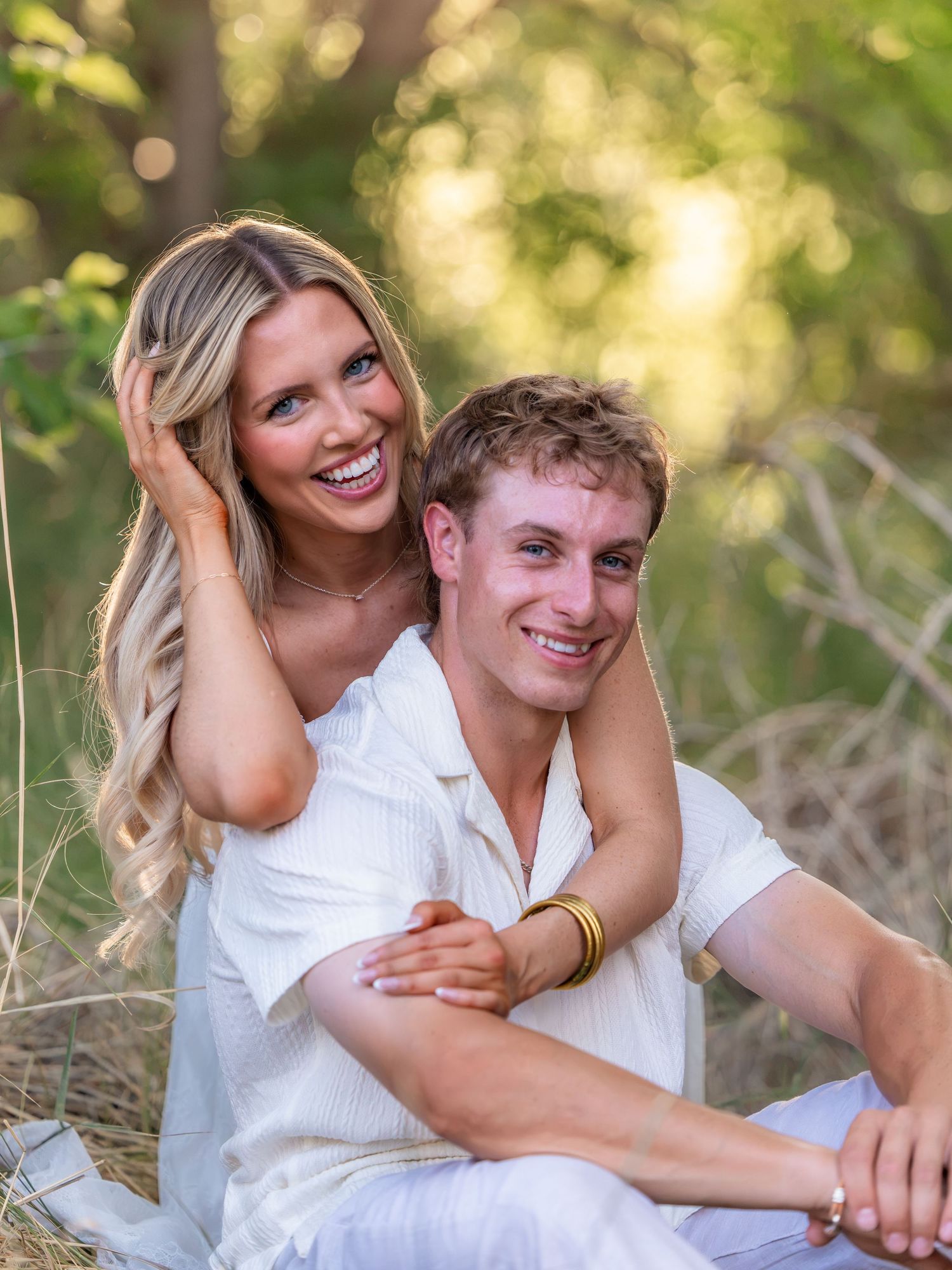 A smiling couple in white outfits poses outdoors in a sunny field with green trees in the background.