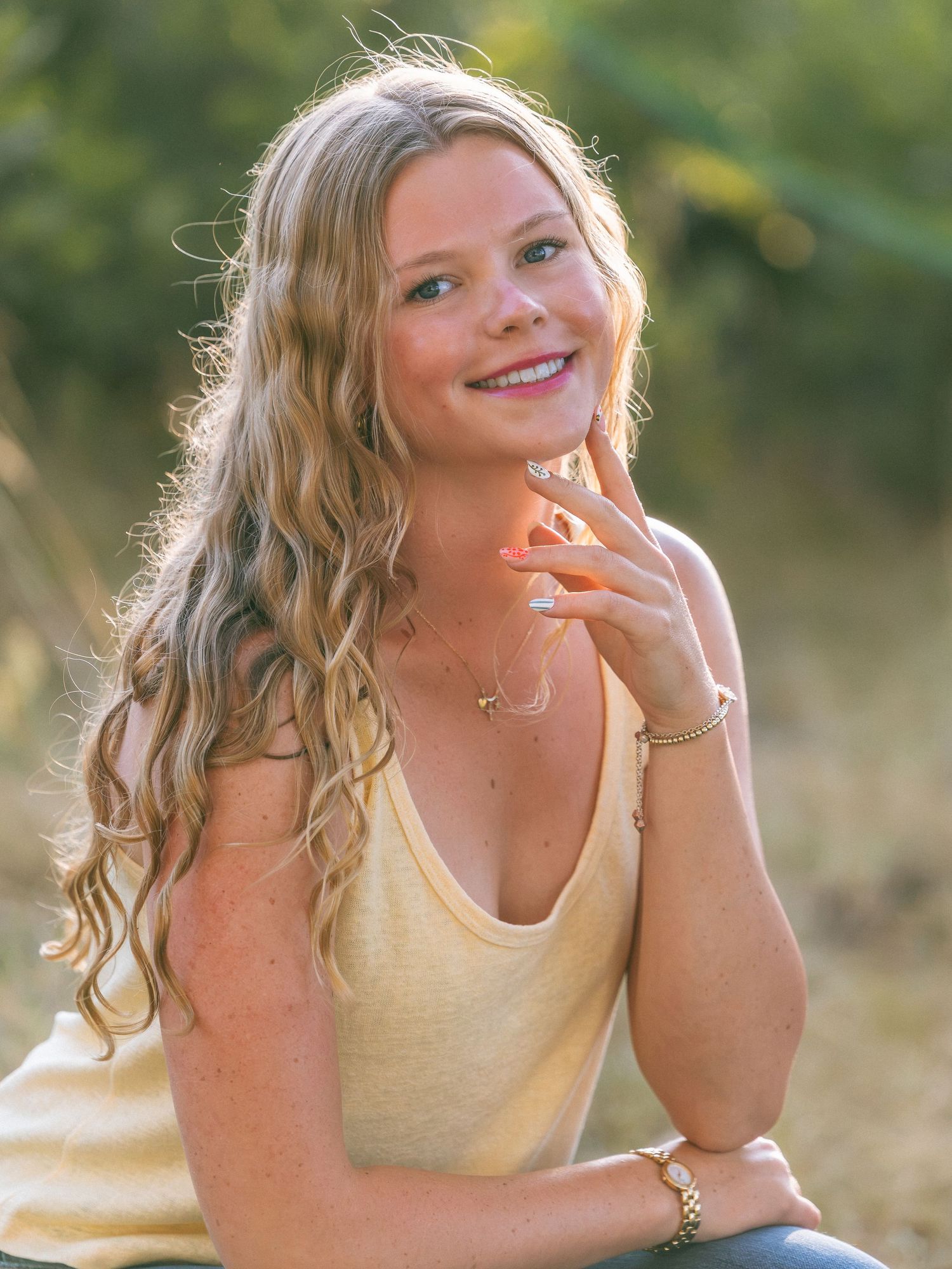 Young blonde woman with wavy hair smiling outdoors, wearing a yellow tank top and gold jewelry in warm sunlight.