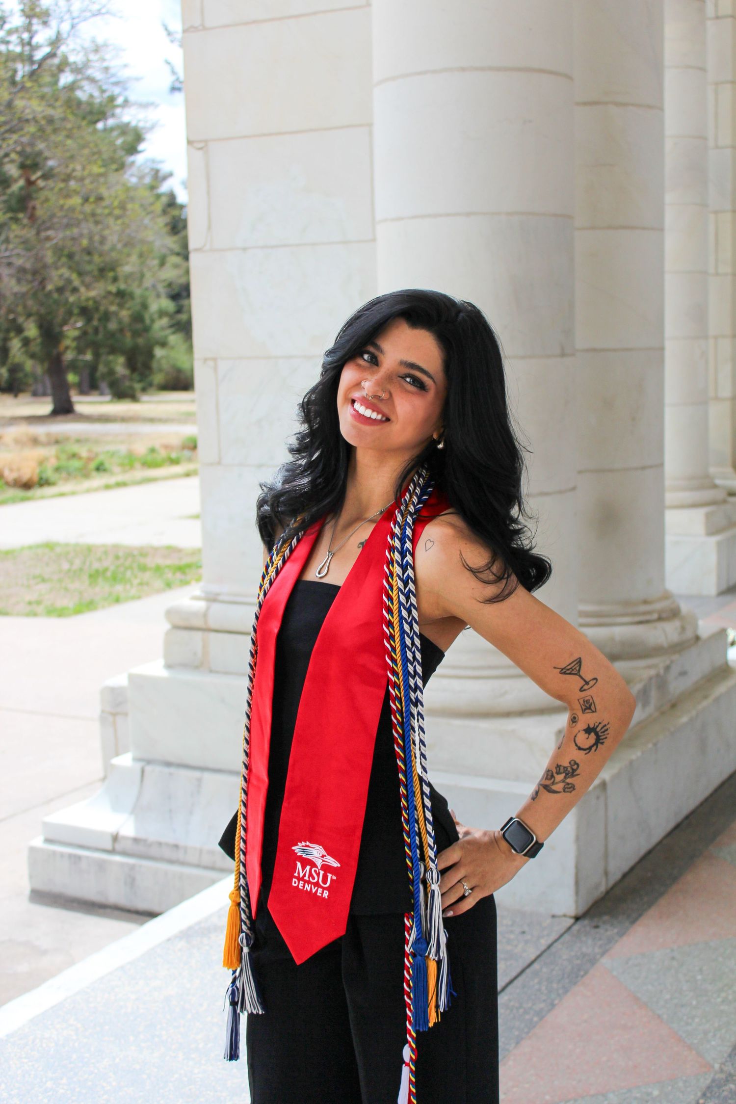 Smiling graduate poses in black outfit with red MSU stole and honor cords between white columns outdoors.