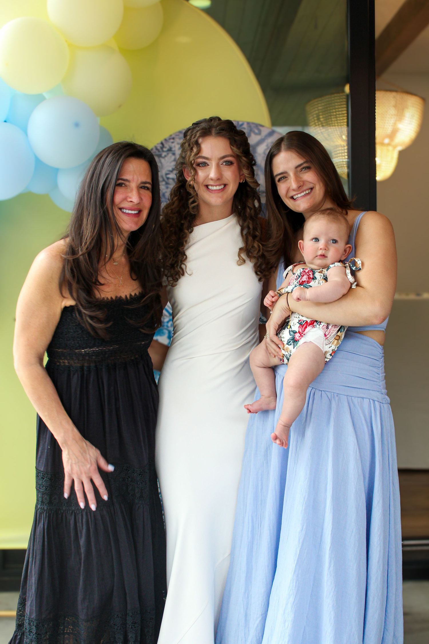 Three women and a baby pose together at a celebration with colorful balloon arch backdrop.