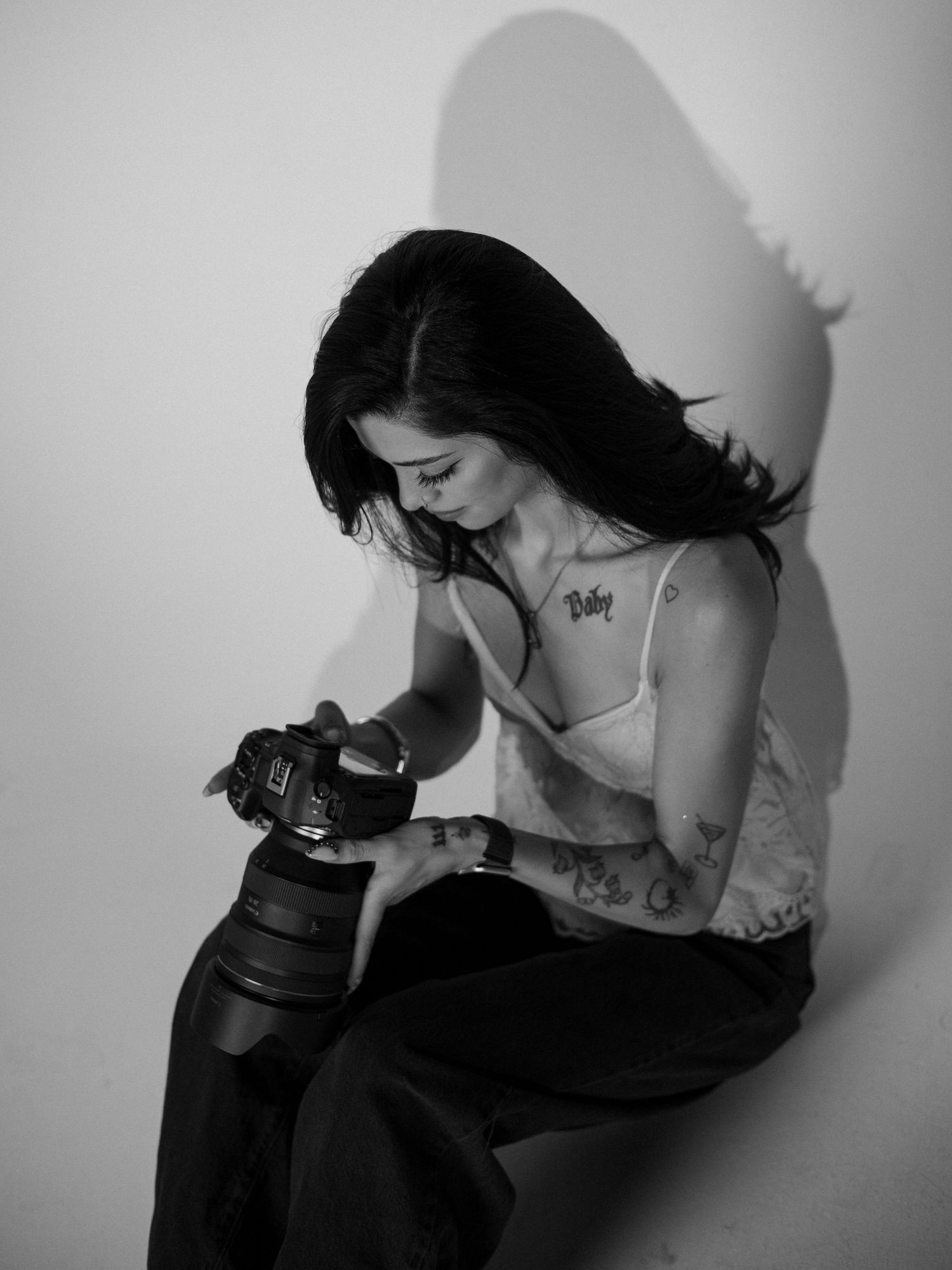 Black and white photo of a tattooed woman sitting and examining a DSLR camera, casting a dramatic shadow.