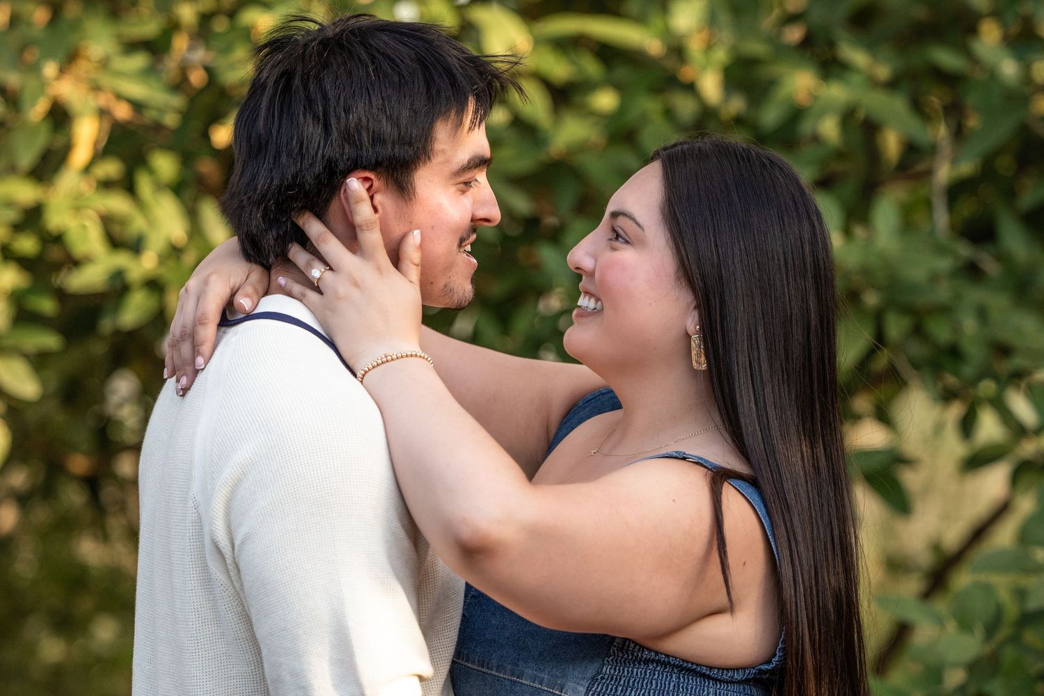 A smiling couple gazes at each other outdoors, the woman holding the man's face tenderly among green foliage.