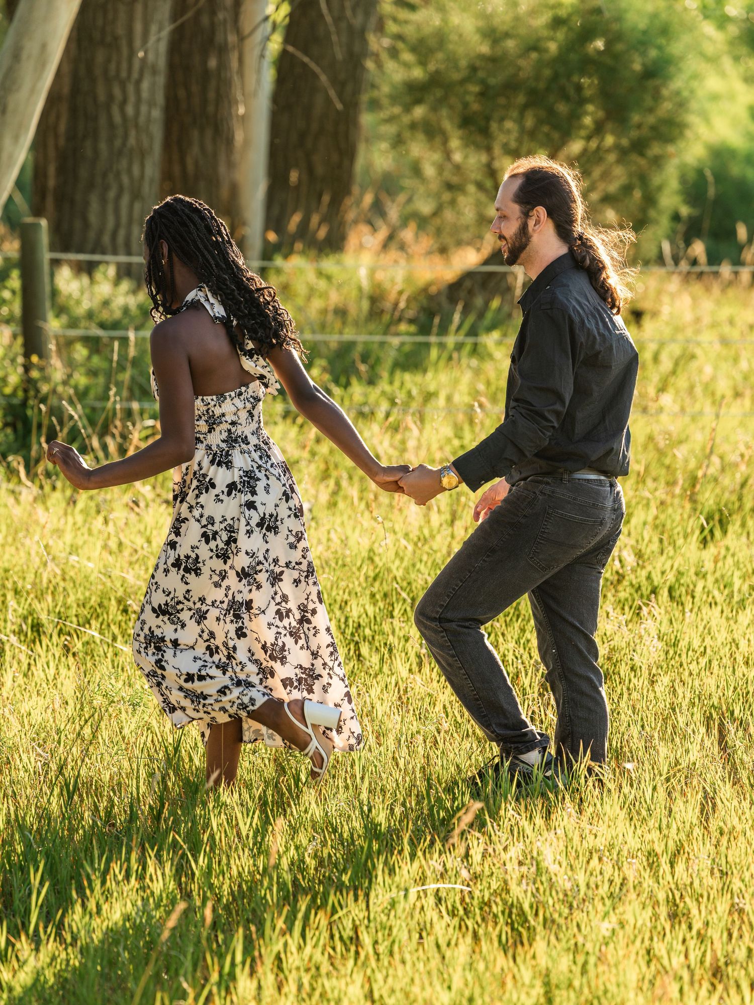 A joyful interracial couple holds hands and dances through a sunlit green meadow, the woman in a floral dress.