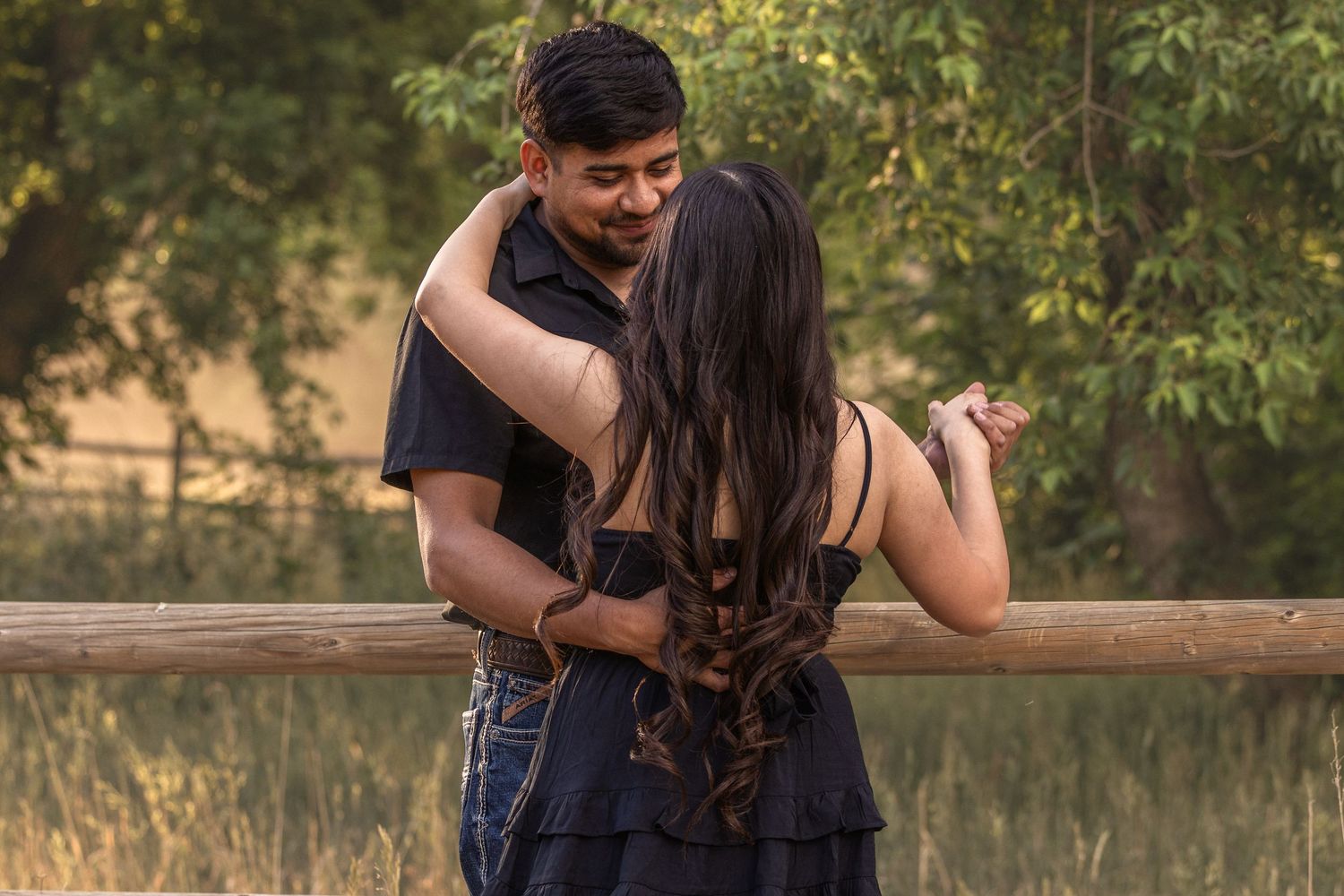 Romantic couple embracing and smiling at each other near a rustic wooden fence in a sunny outdoor setting.