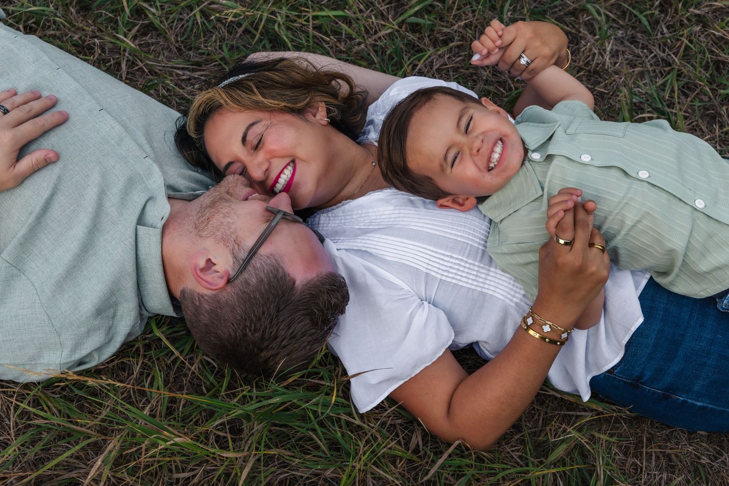 A joyful family of three lying in grass, laughing together in a candid outdoor portrait.