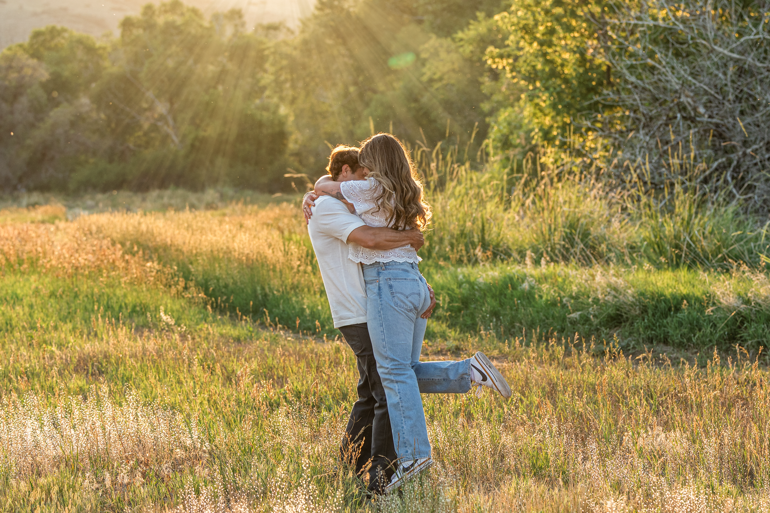 Couple embracing in a sunlit meadow with golden hour light, surrounded by green grass and trees.