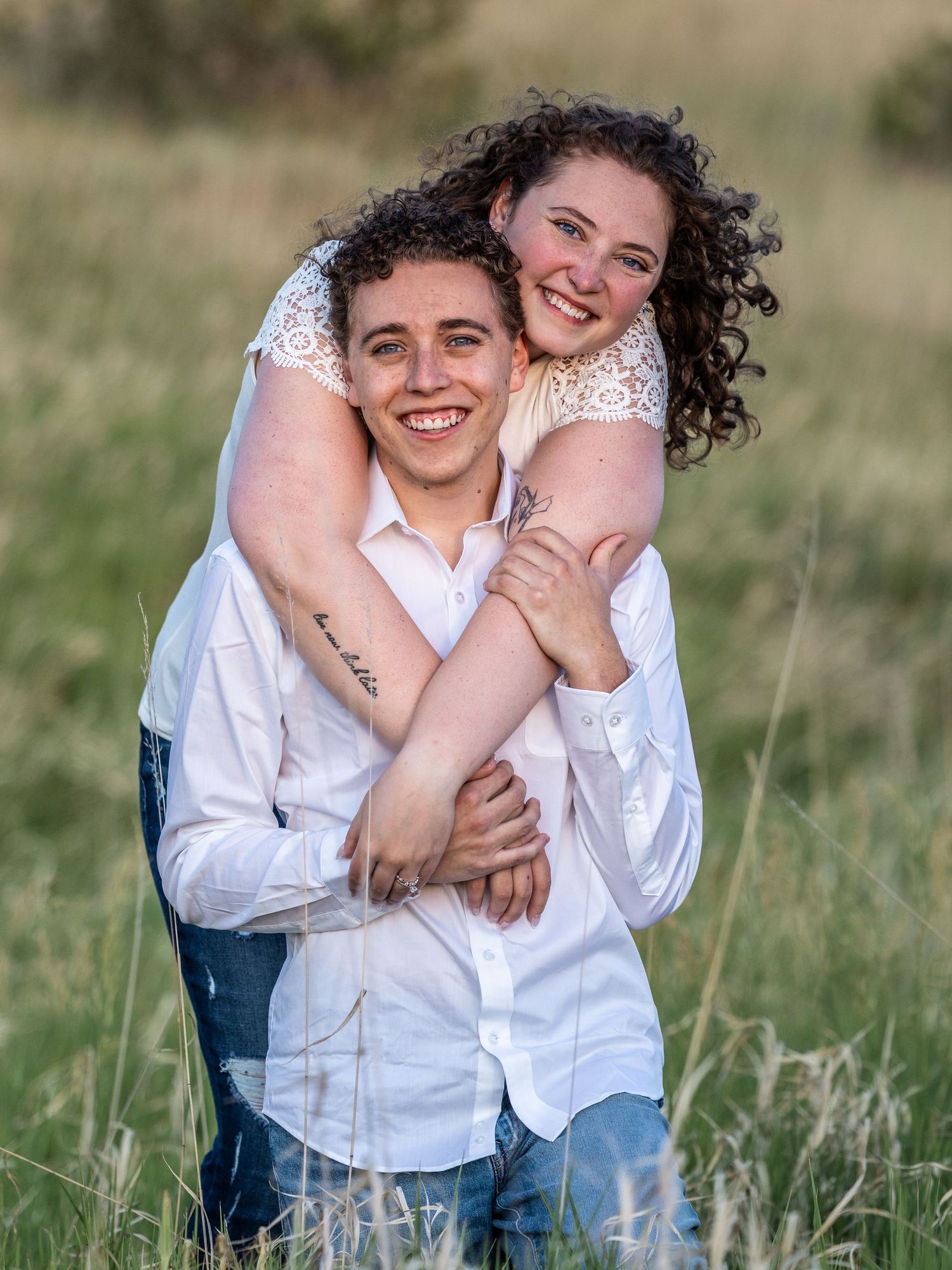 A smiling couple in white shirts embraces in a green field, the woman hugging the man from behind in a joyful pose.