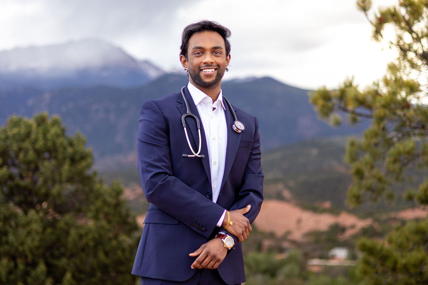 Doctor in navy suit with stethoscope smiling outdoors with mountain landscape background.