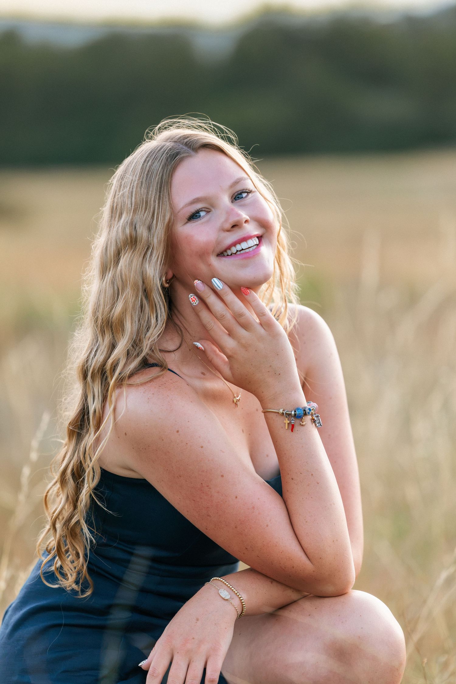 Smiling blonde woman in navy dress posing in golden field, wearing bracelets, hand resting on chin.