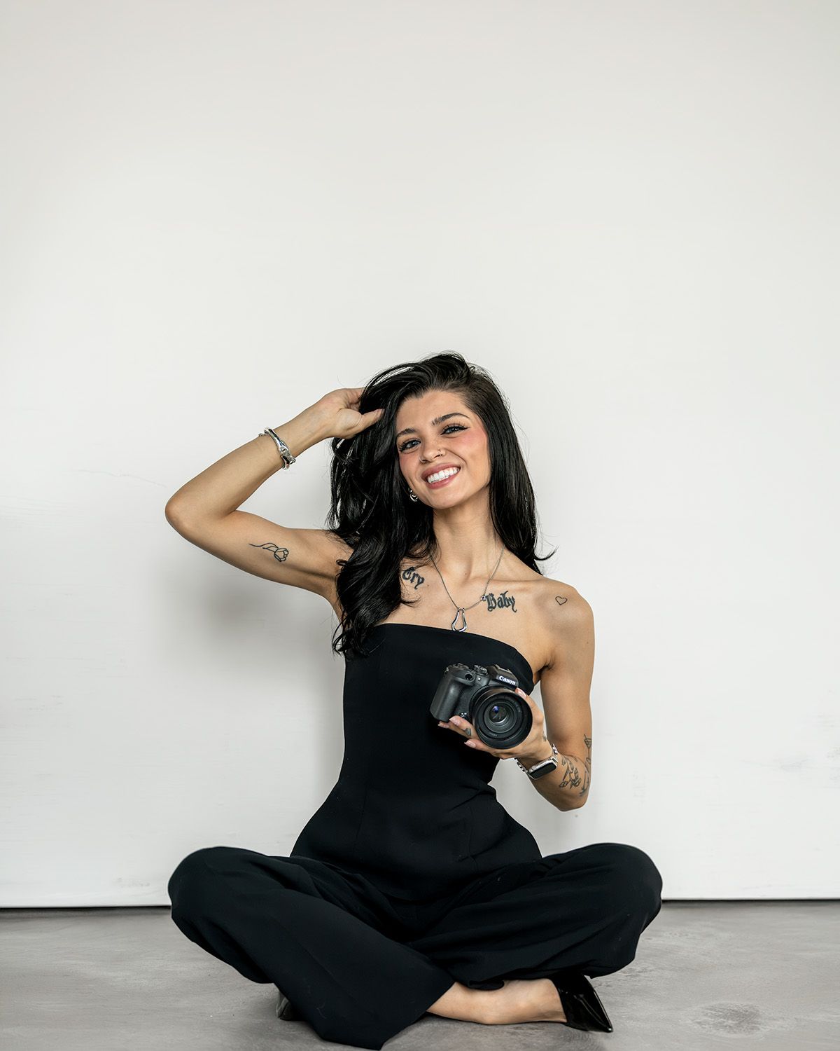 Smiling woman with tattoos in black strapless jumpsuit sitting cross-legged on floor holding a DSLR camera.