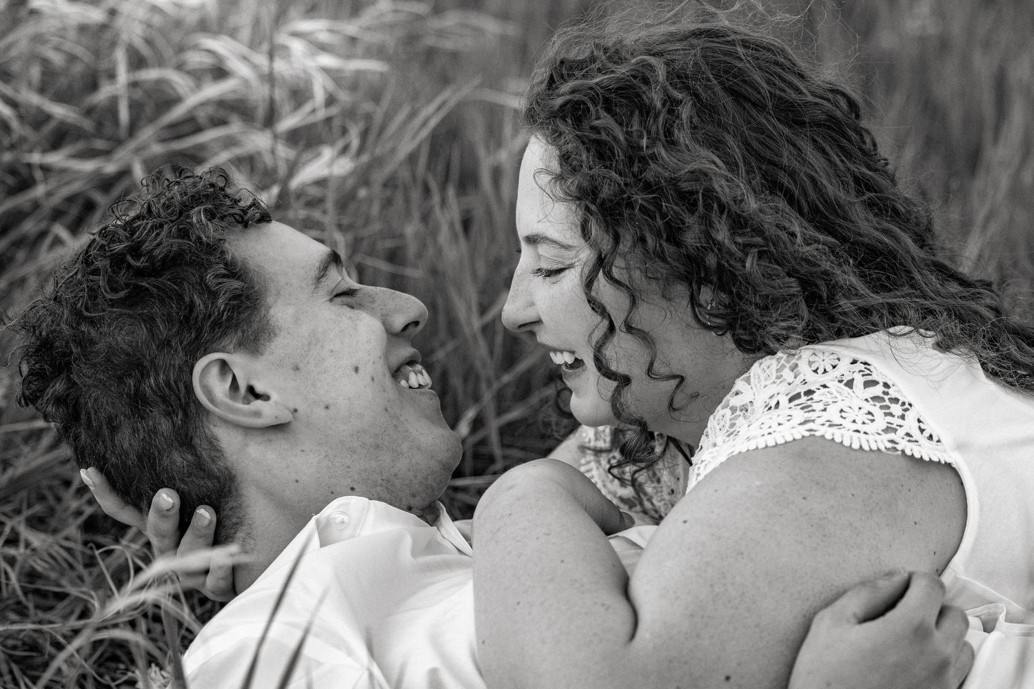 Black and white photo of a romantic couple smiling at each other while embracing outdoors in tall grass.