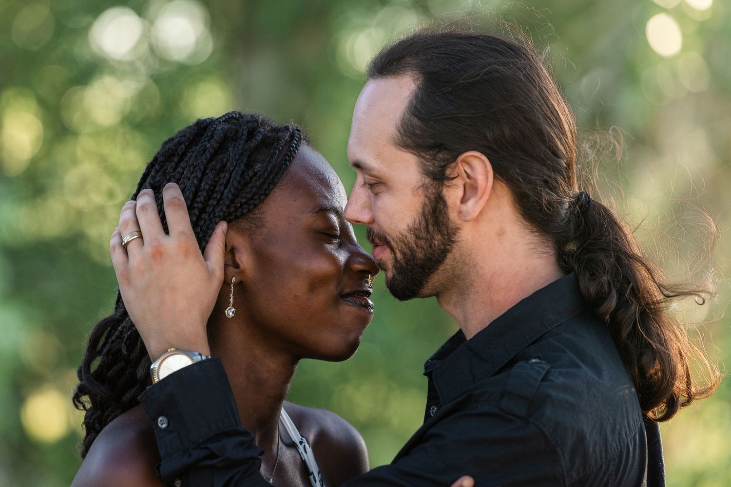 A couple sharing an intimate moment, the woman with braids and the man with long hair, almost kissing outdoors.