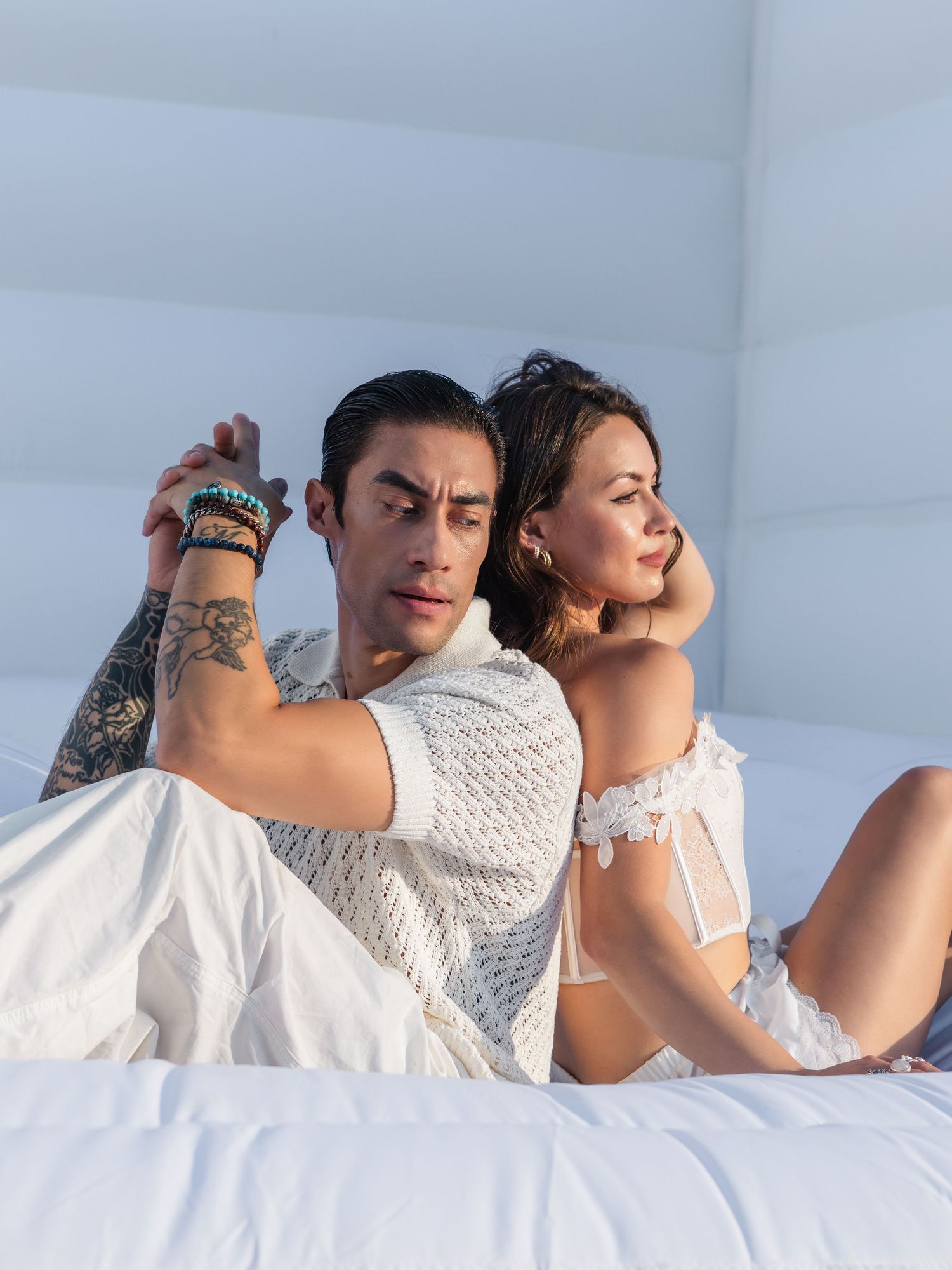 A tattooed man and woman in white outfits sit back-to-back on a white surface in a bright, airy setting.