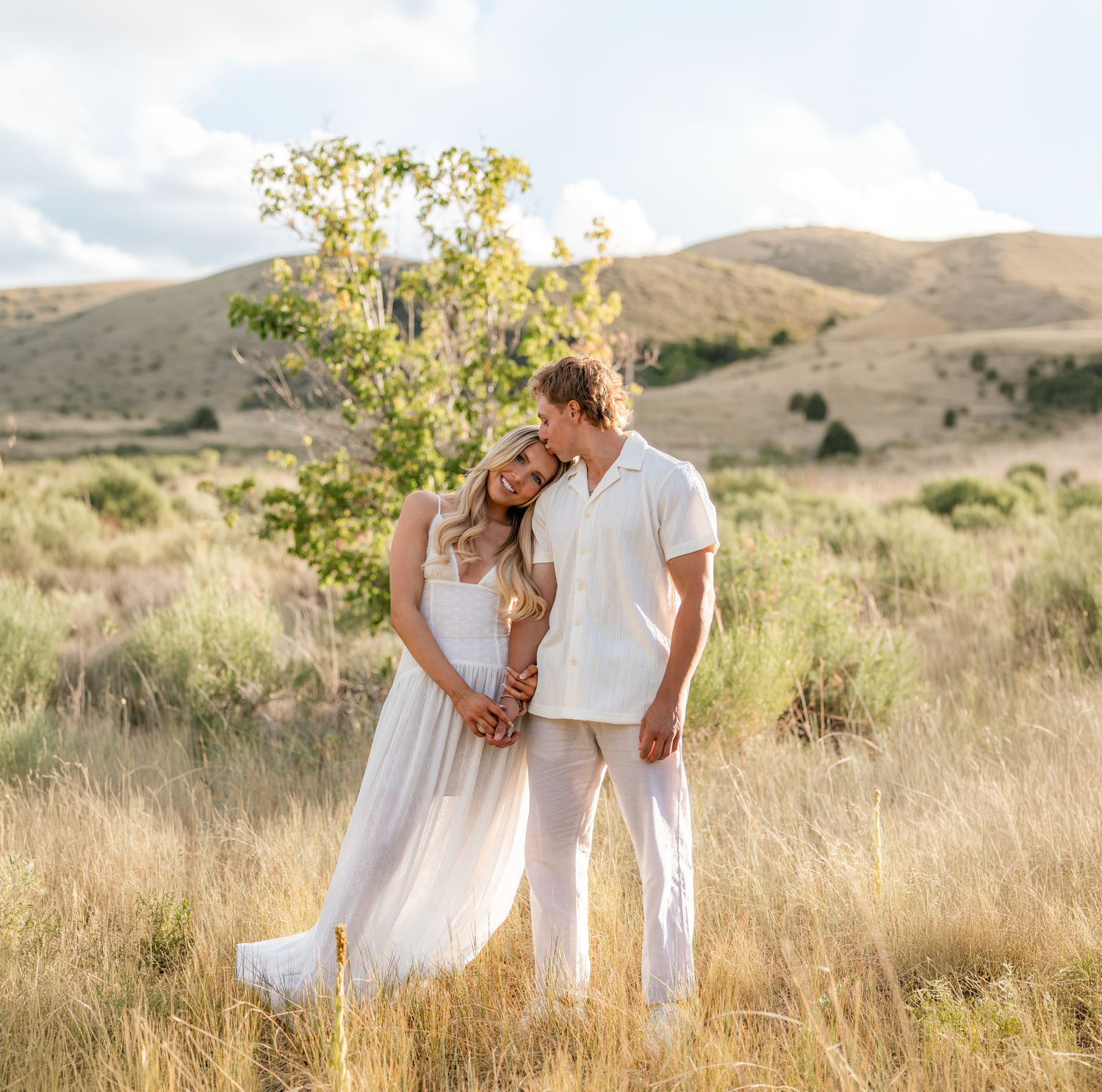 Couple in white outfits embracing in golden field with rolling hills and blue sky backdrop.