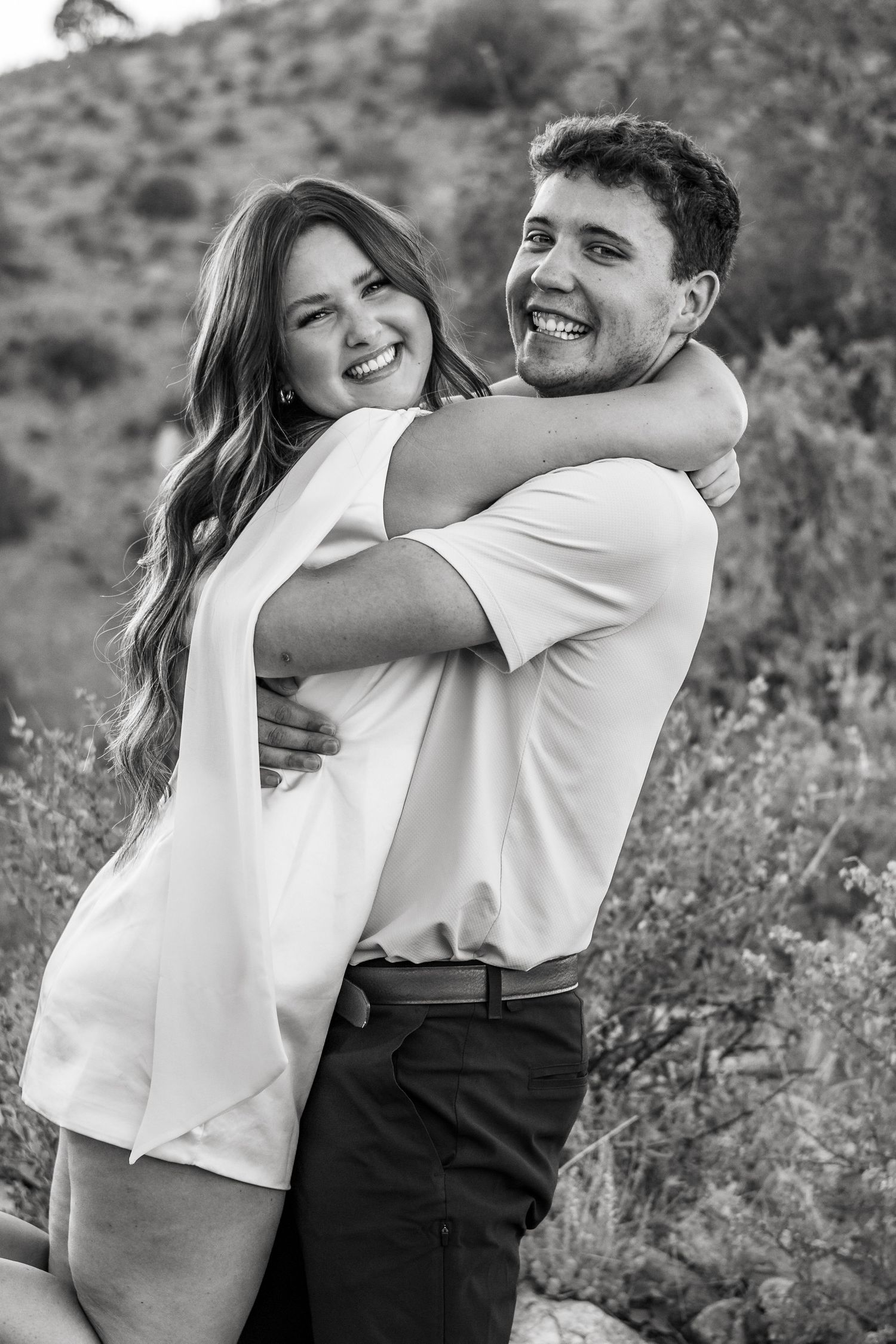 Black and white photo of a smiling couple embracing outdoors with lush greenery in the background.