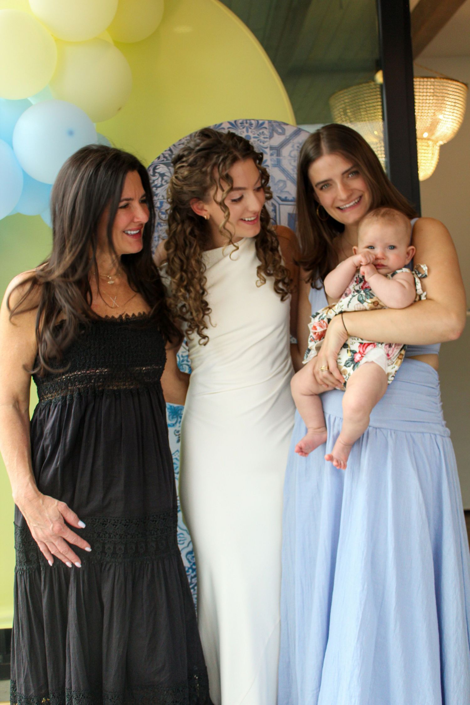 Three women, one holding a baby girl, laugh together at a blue and yellow balloon party.