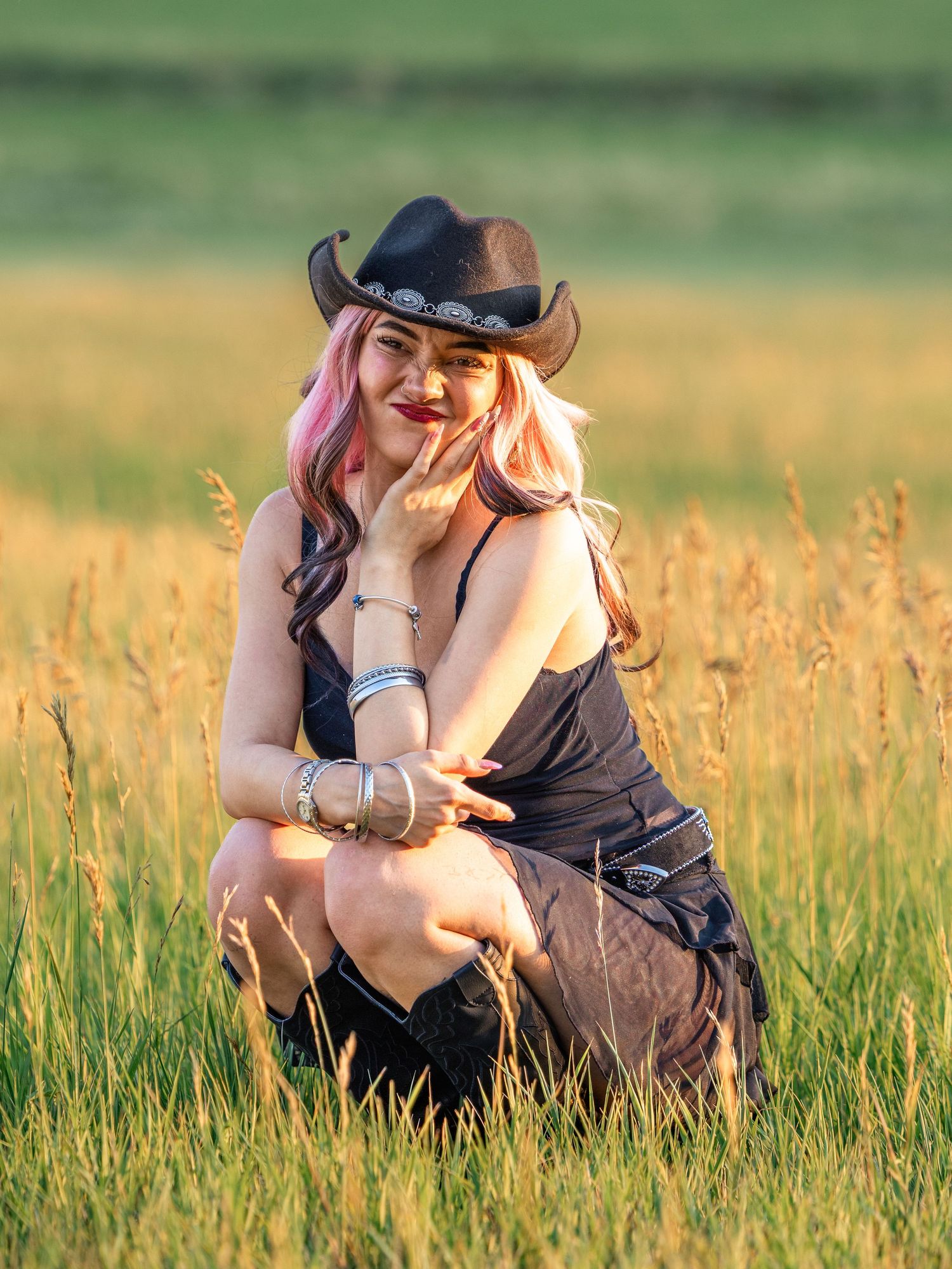 Young woman in black cowboy hat and dress with pink hair crouching in golden sunlit meadow grass.