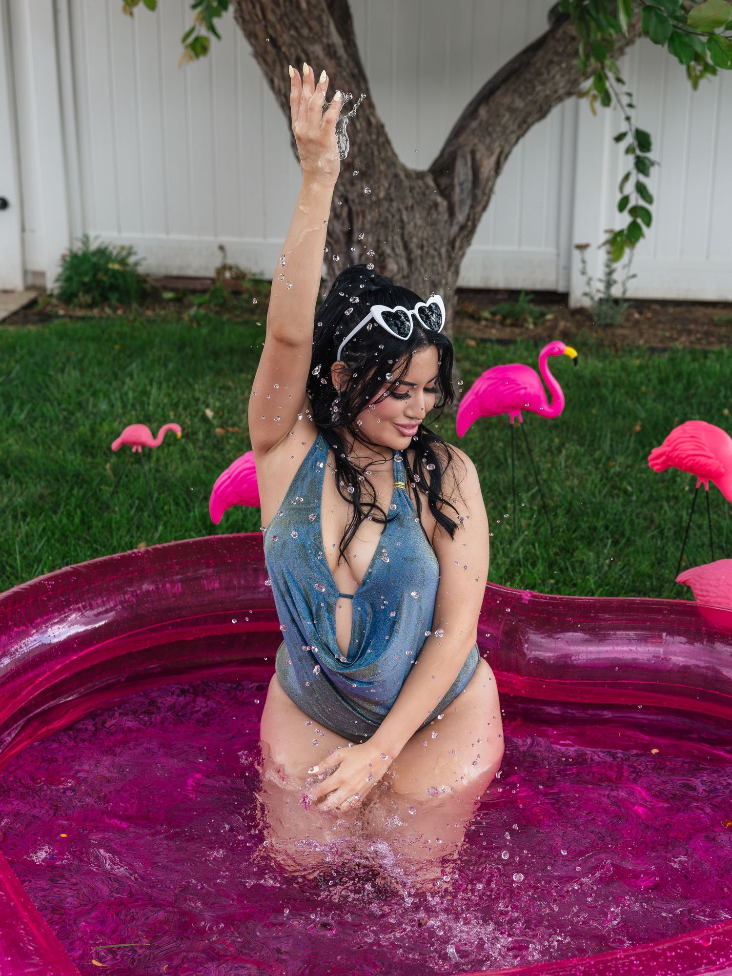 Woman in gray swimsuit splashing in pink inflatable pool outdoors with flamingo decorations and green lawn background.