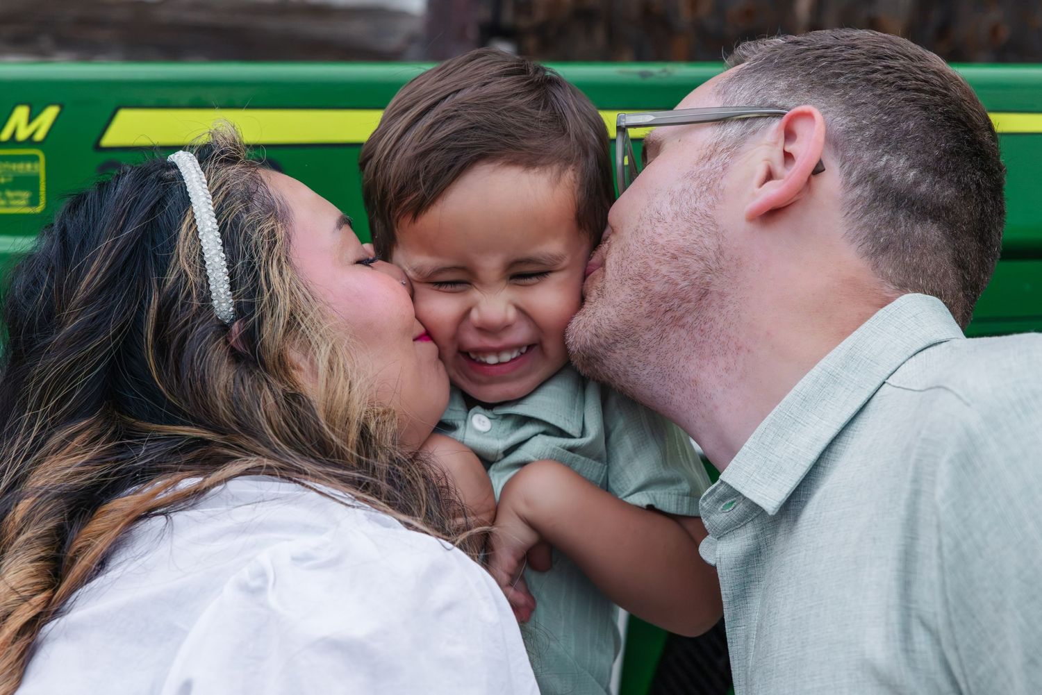 Parents kissing their laughing toddler boy on both cheeks outdoors near a green John Deere tractor.