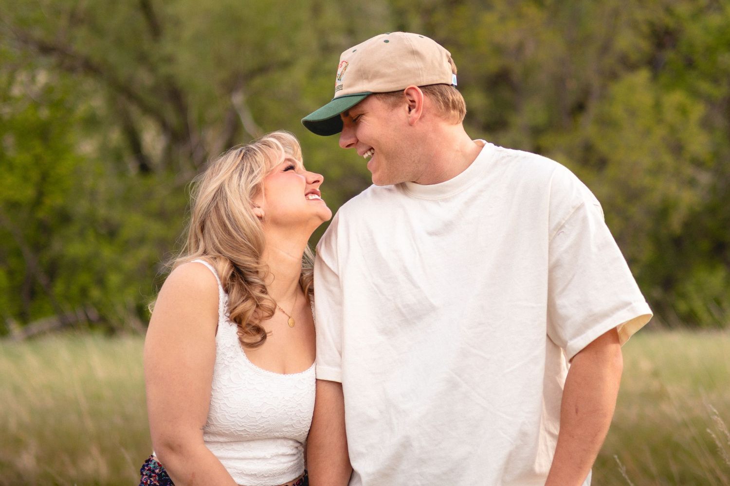 A smiling couple in casual attire share a loving glance outdoors with green trees in the background in Littleton, Colorado.