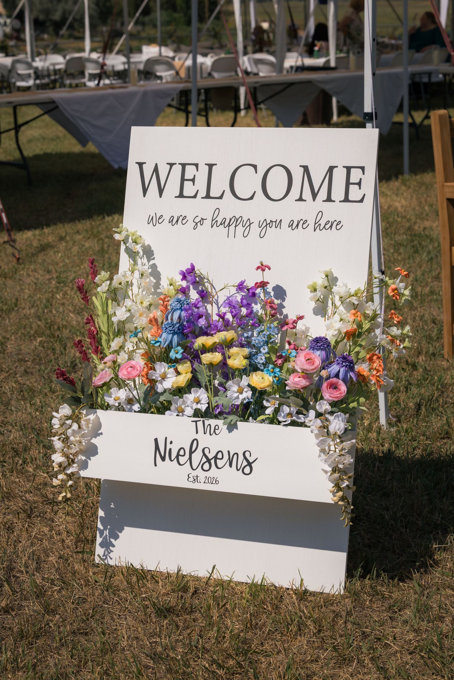 White wedding welcome sign for The Nielsens Est. 2026, adorned with colorful wildflowers, displayed on grass at outdoor ceremony.