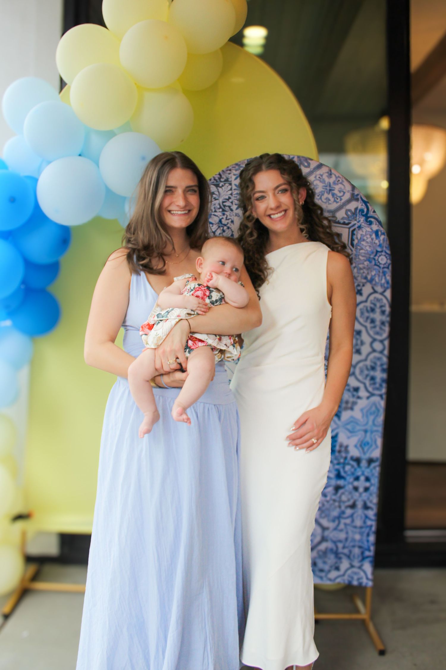 Two women hold a smiling baby at a cheerful event decorated with yellow and blue balloons.