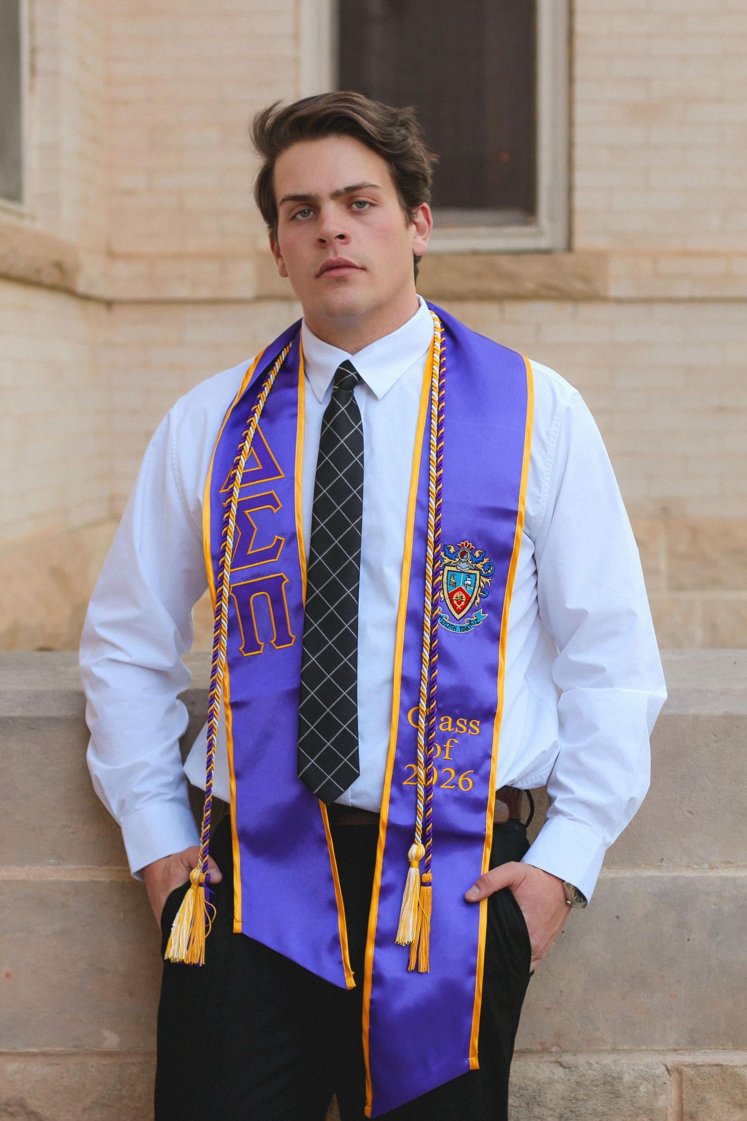 Serious male graduate poses in purple Delta Sigma Pi Class of 2026 stole on stone steps.
