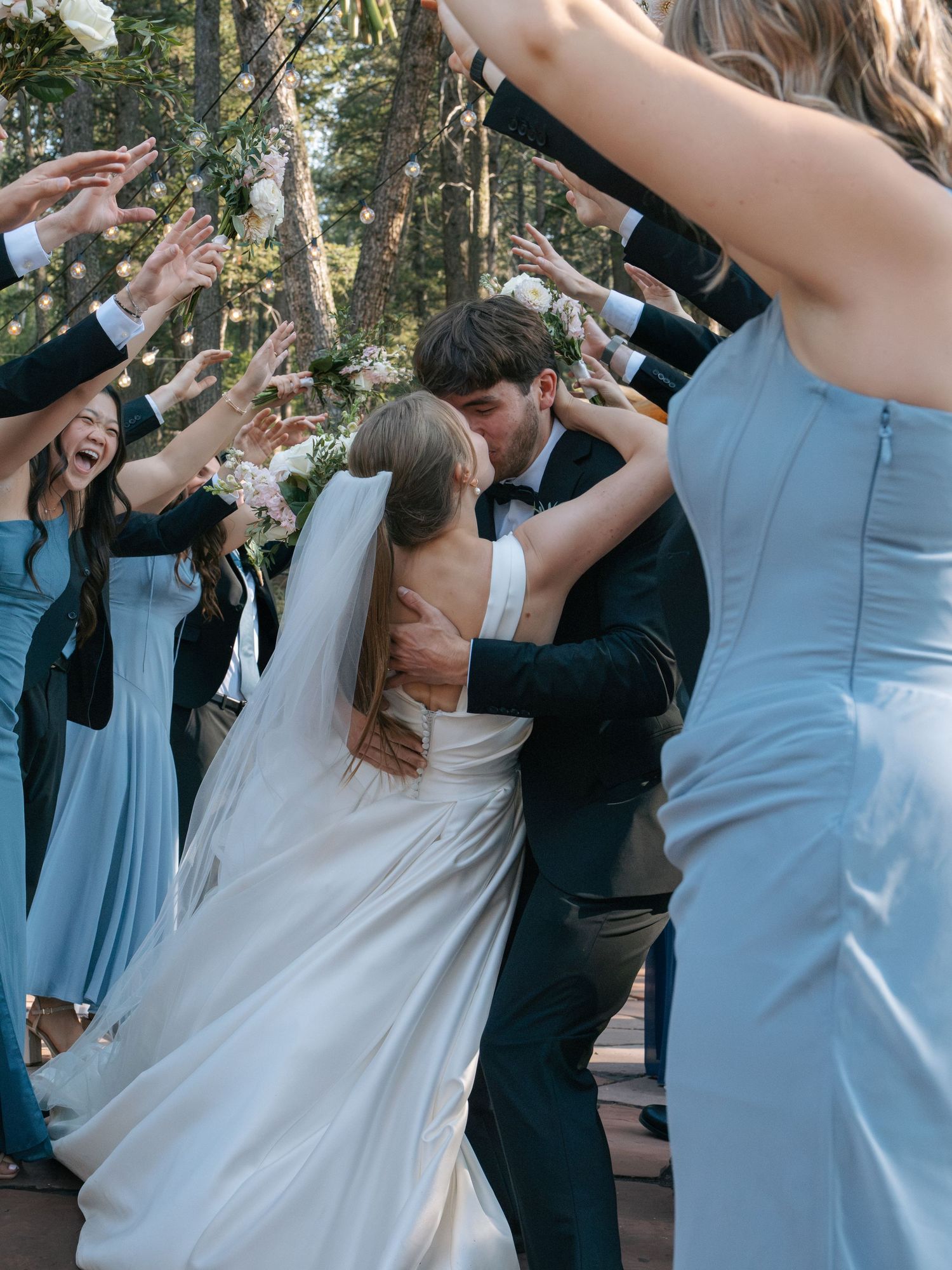 Bride and groom share a kiss while wedding party cheers with raised arms in a forest ceremony.