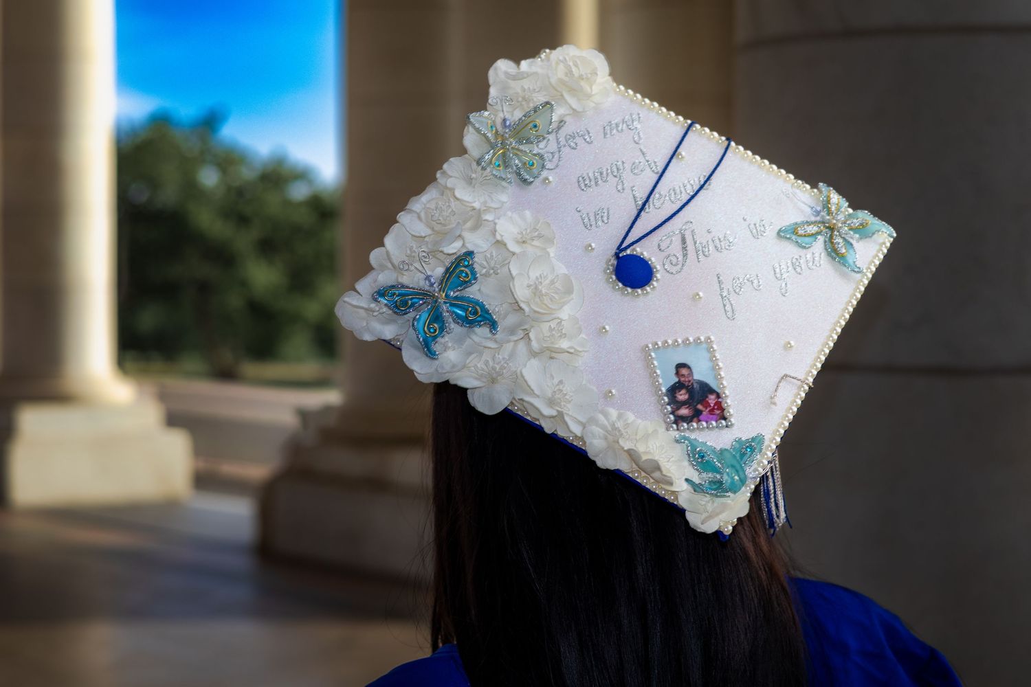 Decorated graduation cap with butterflies, flowers, and photos on a graduate in blue regalia outdoors.