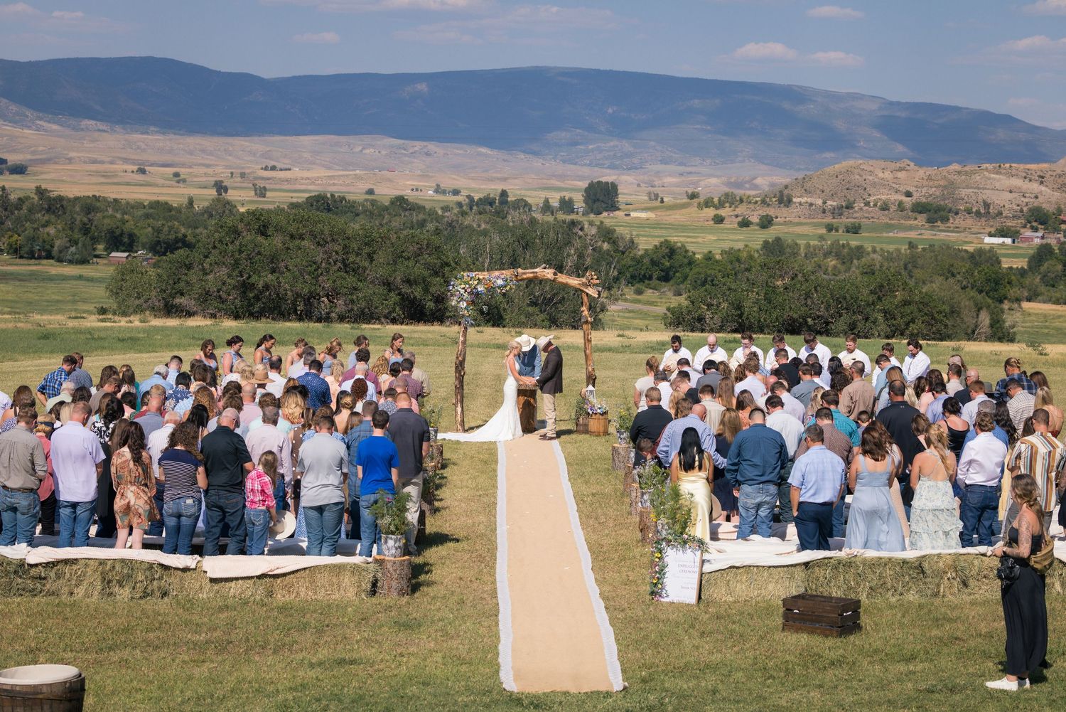 Outdoor wedding ceremony in a scenic mountain valley with guests seated on both sides of a burlap aisle runner.
