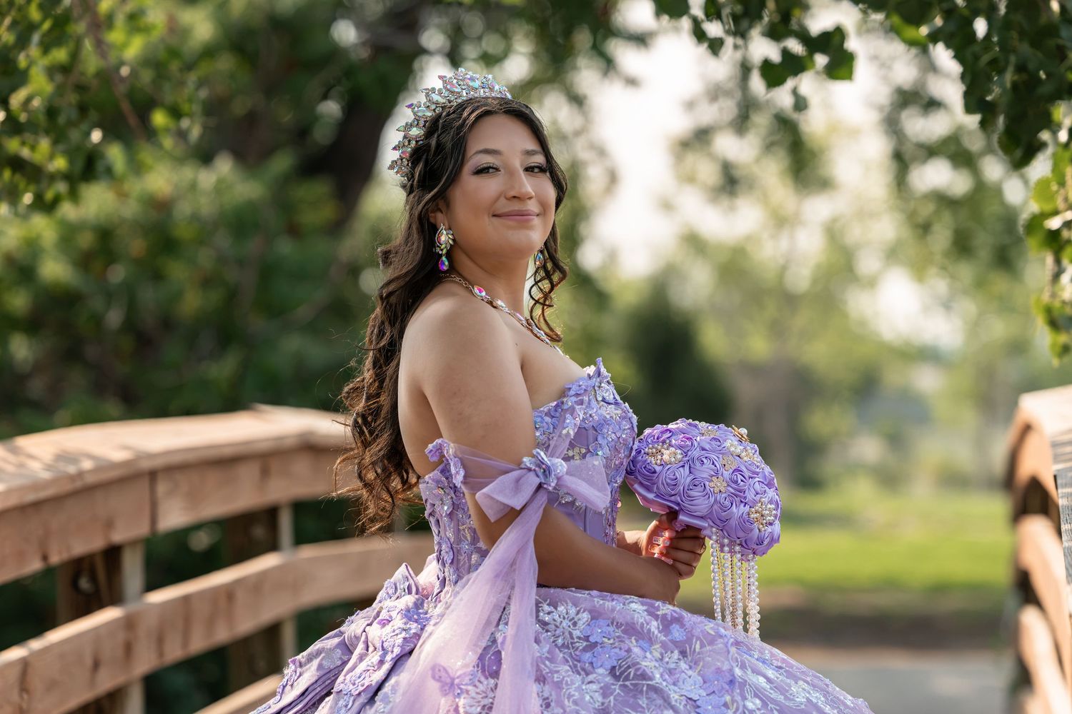Young woman in lavender quinceañera ball gown with floral bouquet and tiara poses on wooden bridge outdoors.