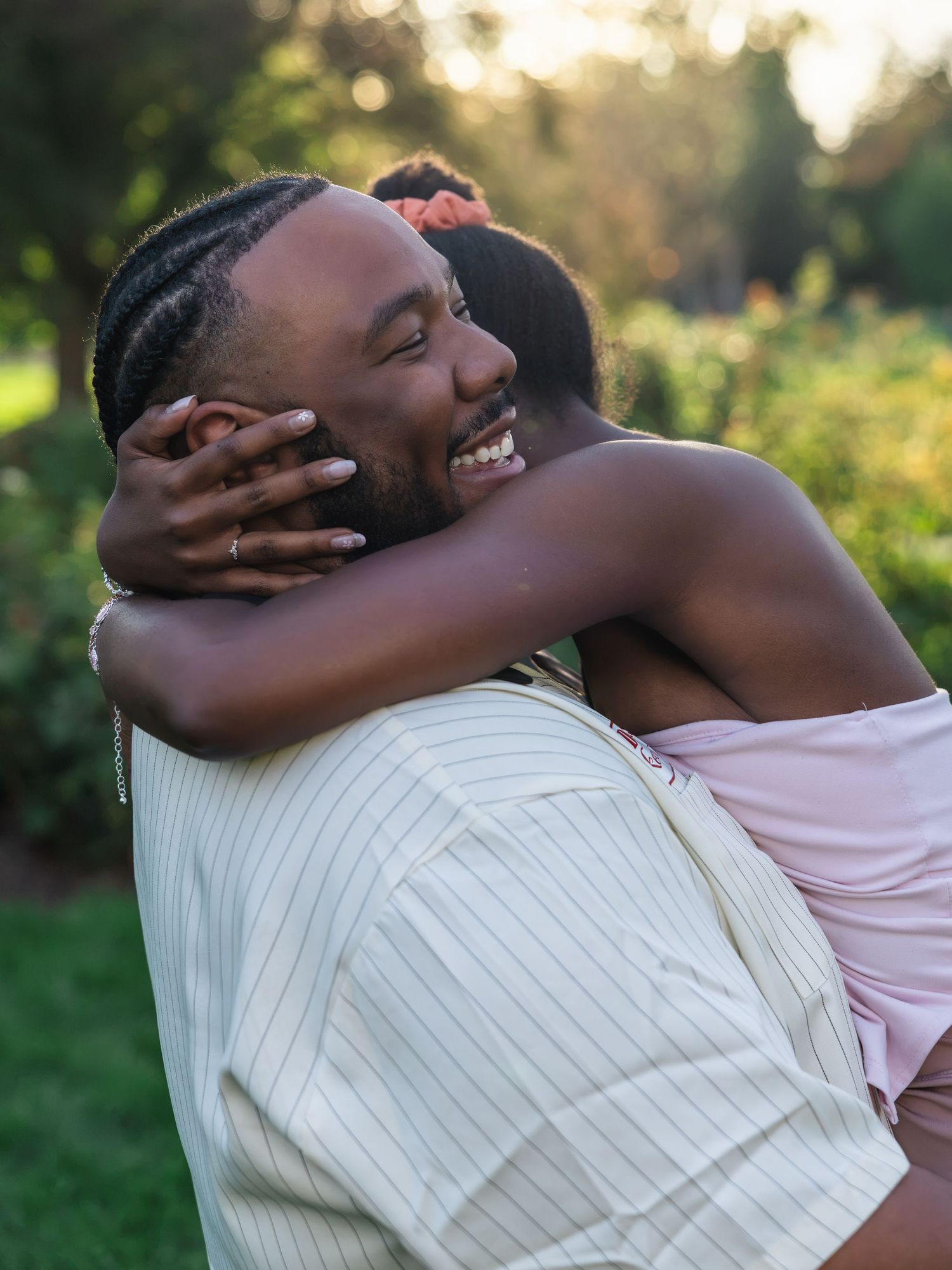 A joyful man with braids hugs a woman in a pink dress outdoors at sunset in a lush green garden.