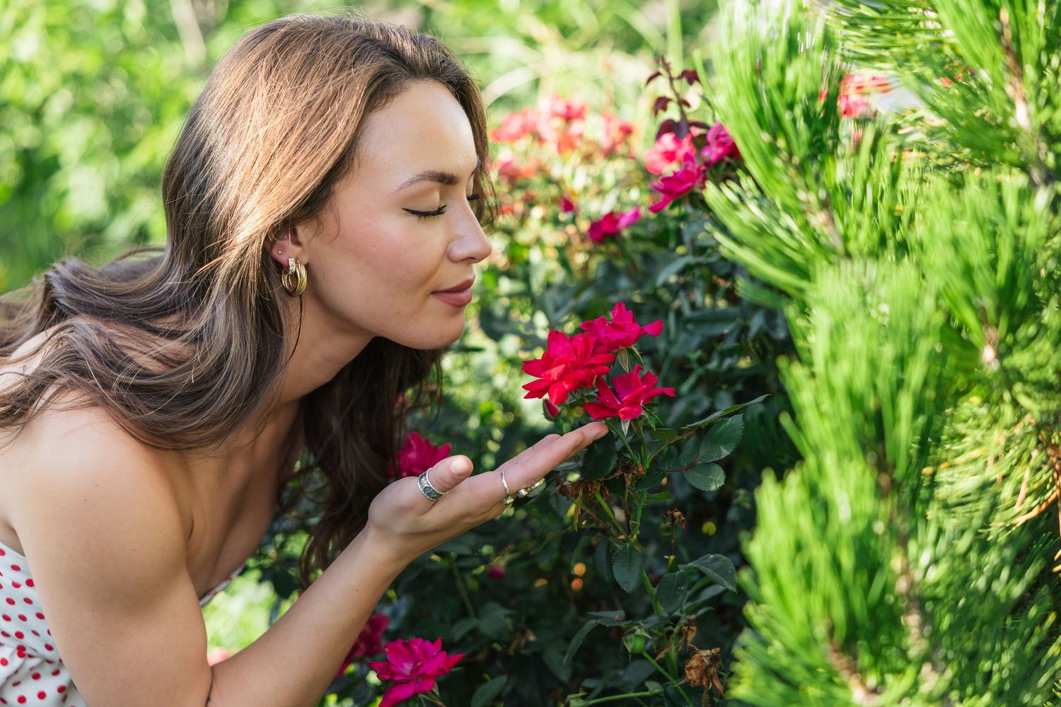Woman with long brown hair smelling red roses in a lush green garden on a sunny day.