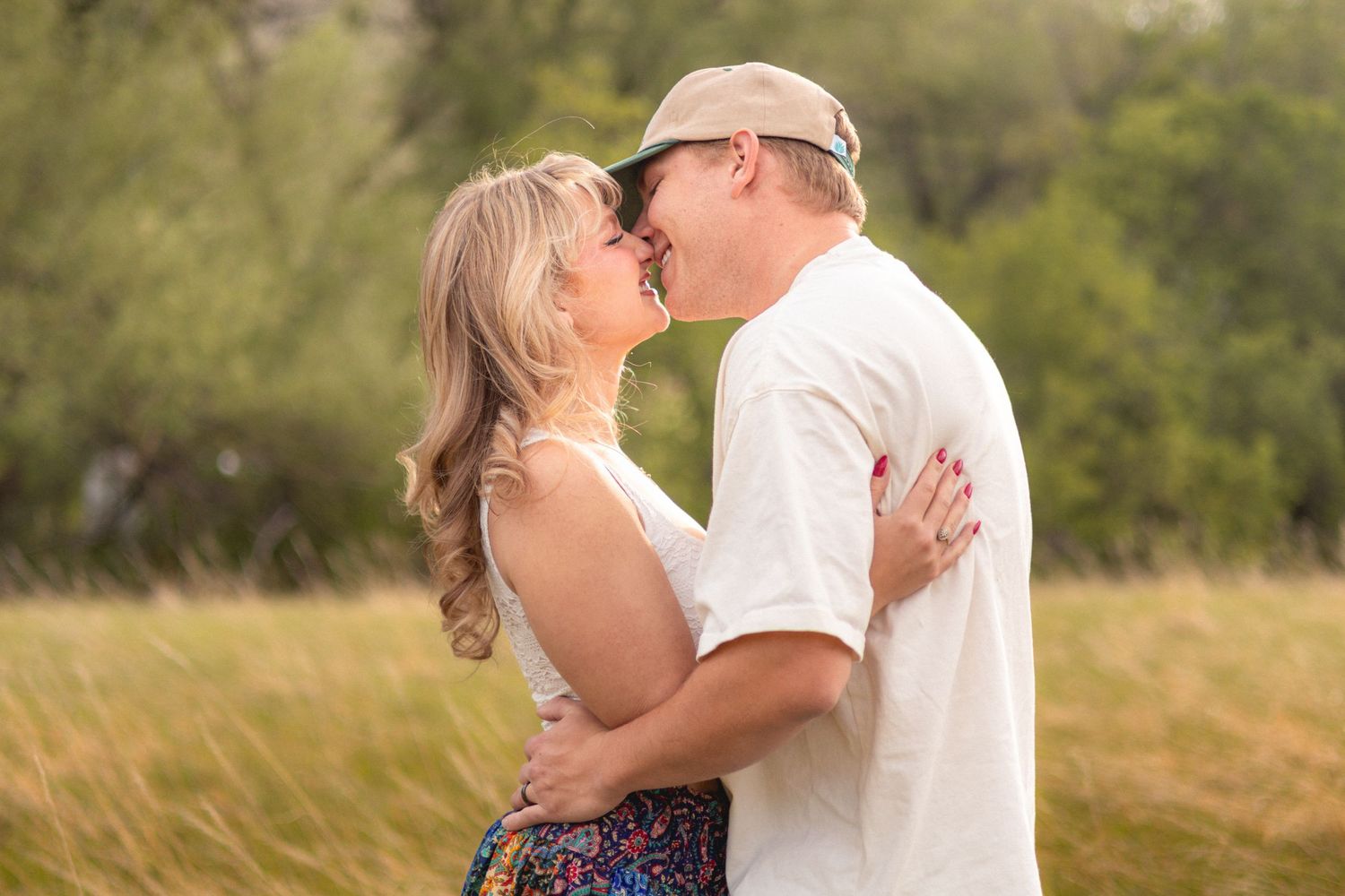 A couple shares a romantic kiss in a lush green field, the man wearing a cap and white shirt embracing a blonde woman.